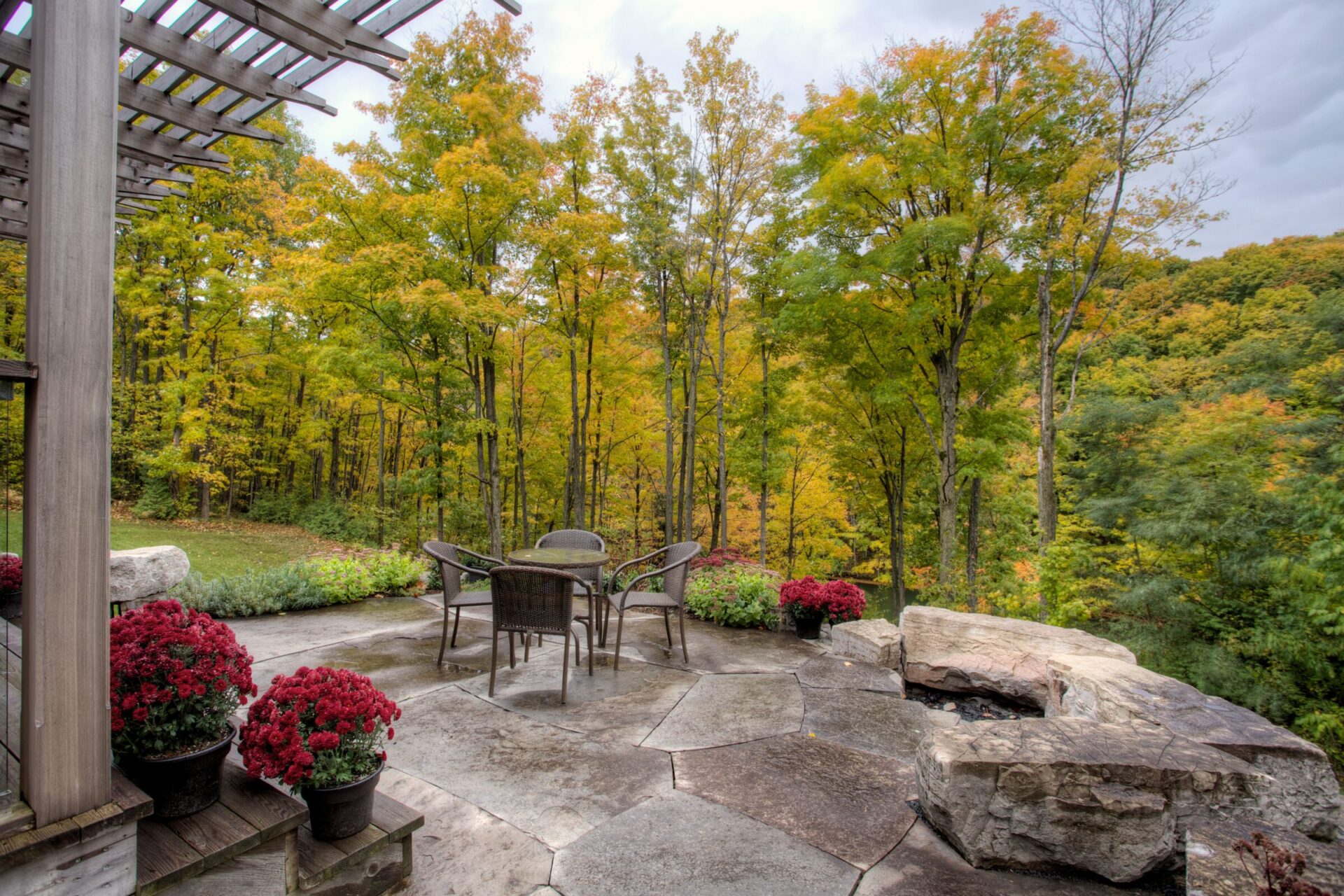 A serene patio features chairs, a table, and red flowers, surrounded by vibrant autumn trees, creating a peaceful outdoor setting.