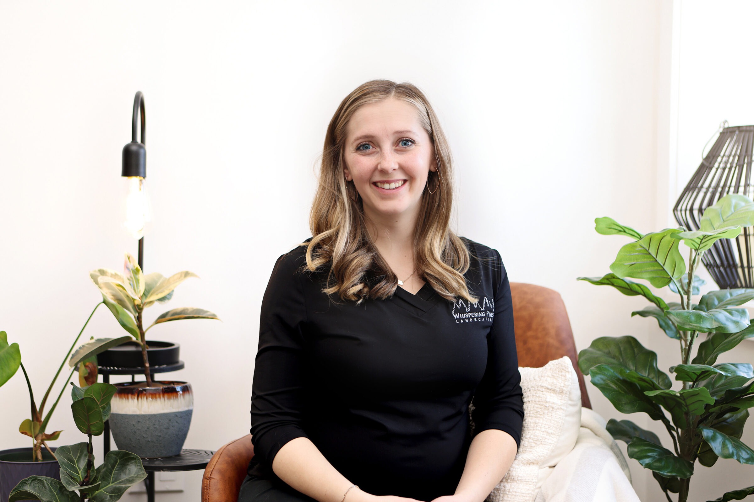 A person sitting in a cozy, plant-decorated room. Wearing a black shirt, smiling, with a modern lamp and potted plants beside them.