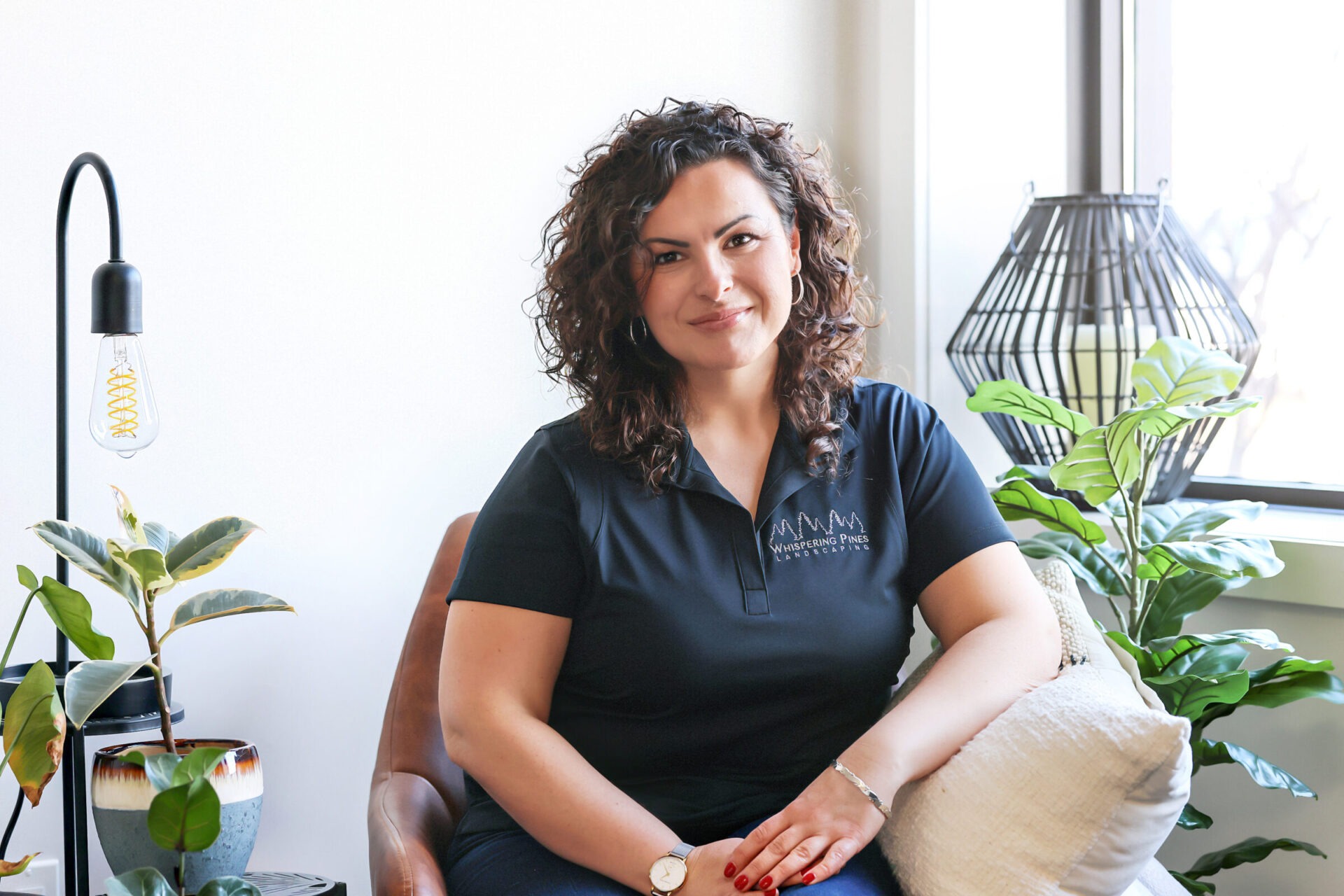 A person with curly hair is seated, smiling, and surrounded by plants in a well-lit room with modern decor.