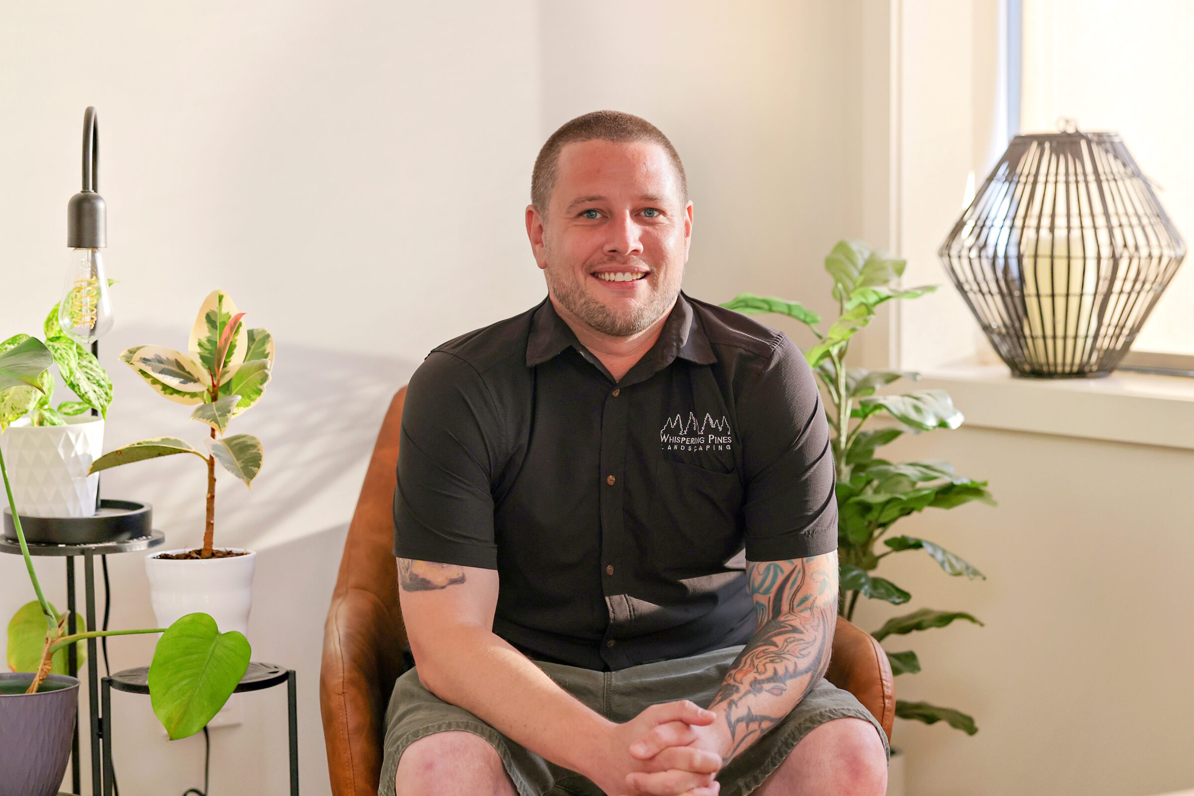 A smiling person sits in a chair surrounded by plants indoors, with a lamp on the right and a warm ambiance.