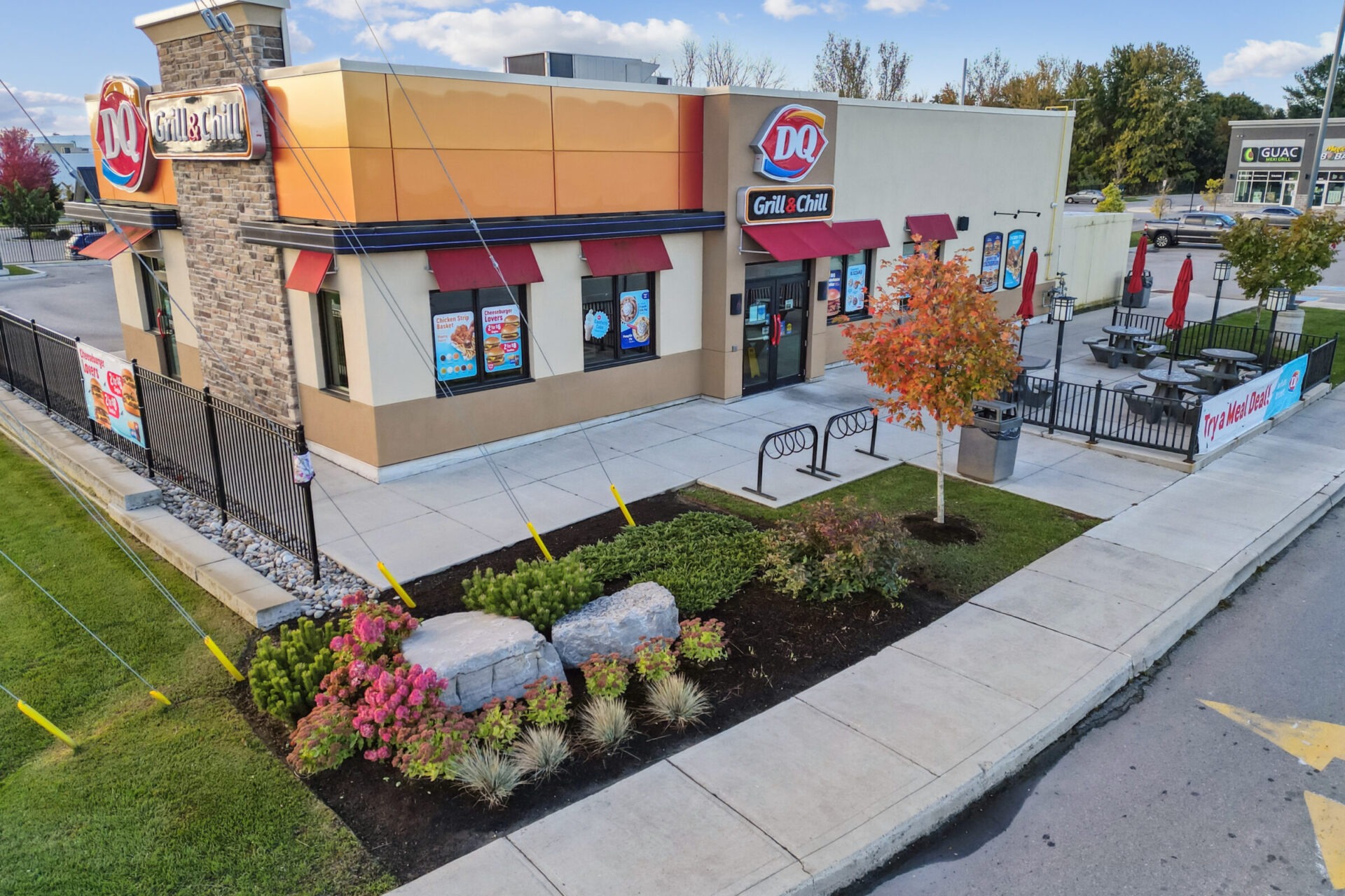 A Dairy Queen Grill & Chill restaurant with outdoor seating, surrounded by greenery and a parking area. Bright signage and autumn-colored tree visible.