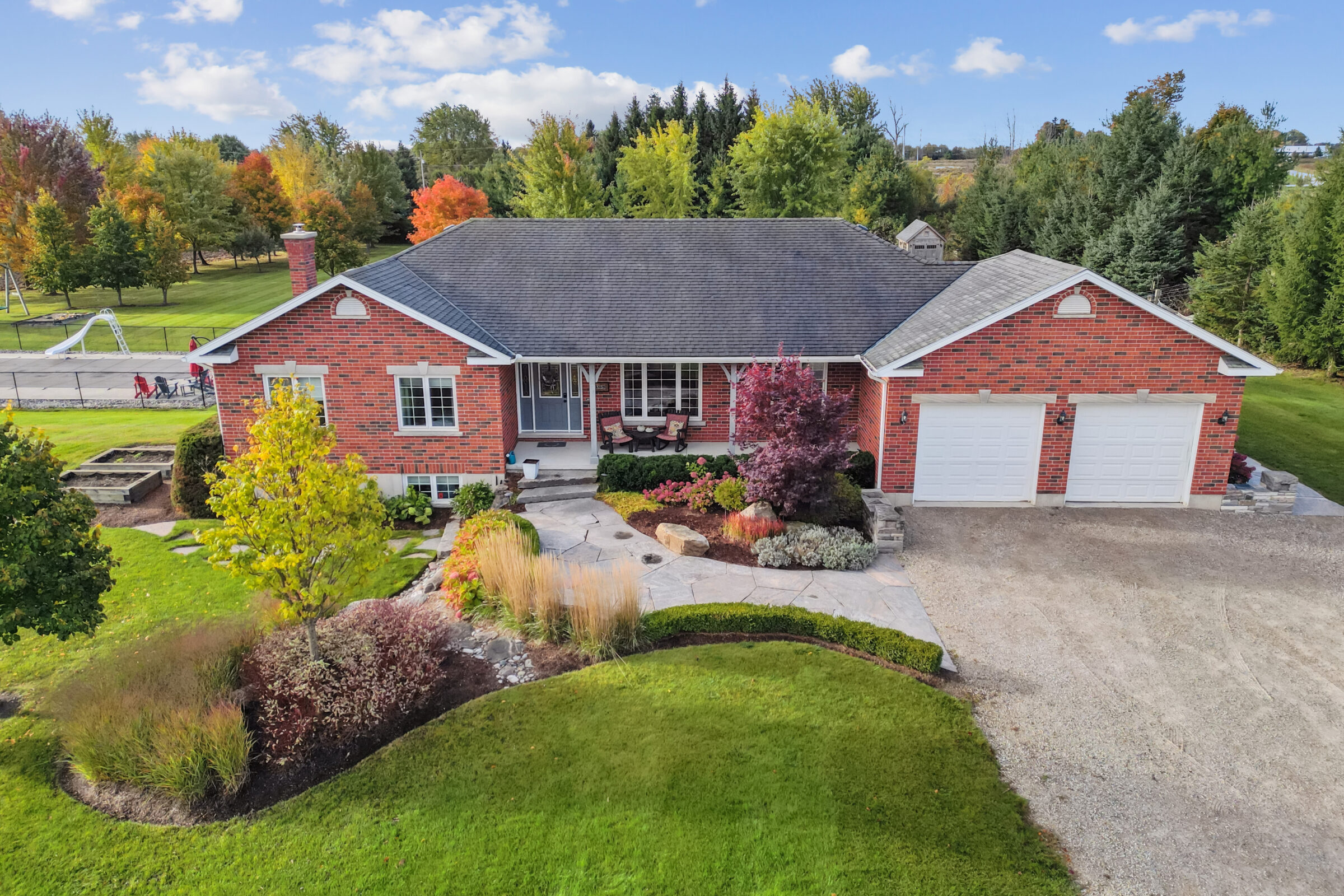 Single-story brick house with manicured lawn, colorful autumn trees, stone path, two-car garage, and adjoining playground visible in the background.
