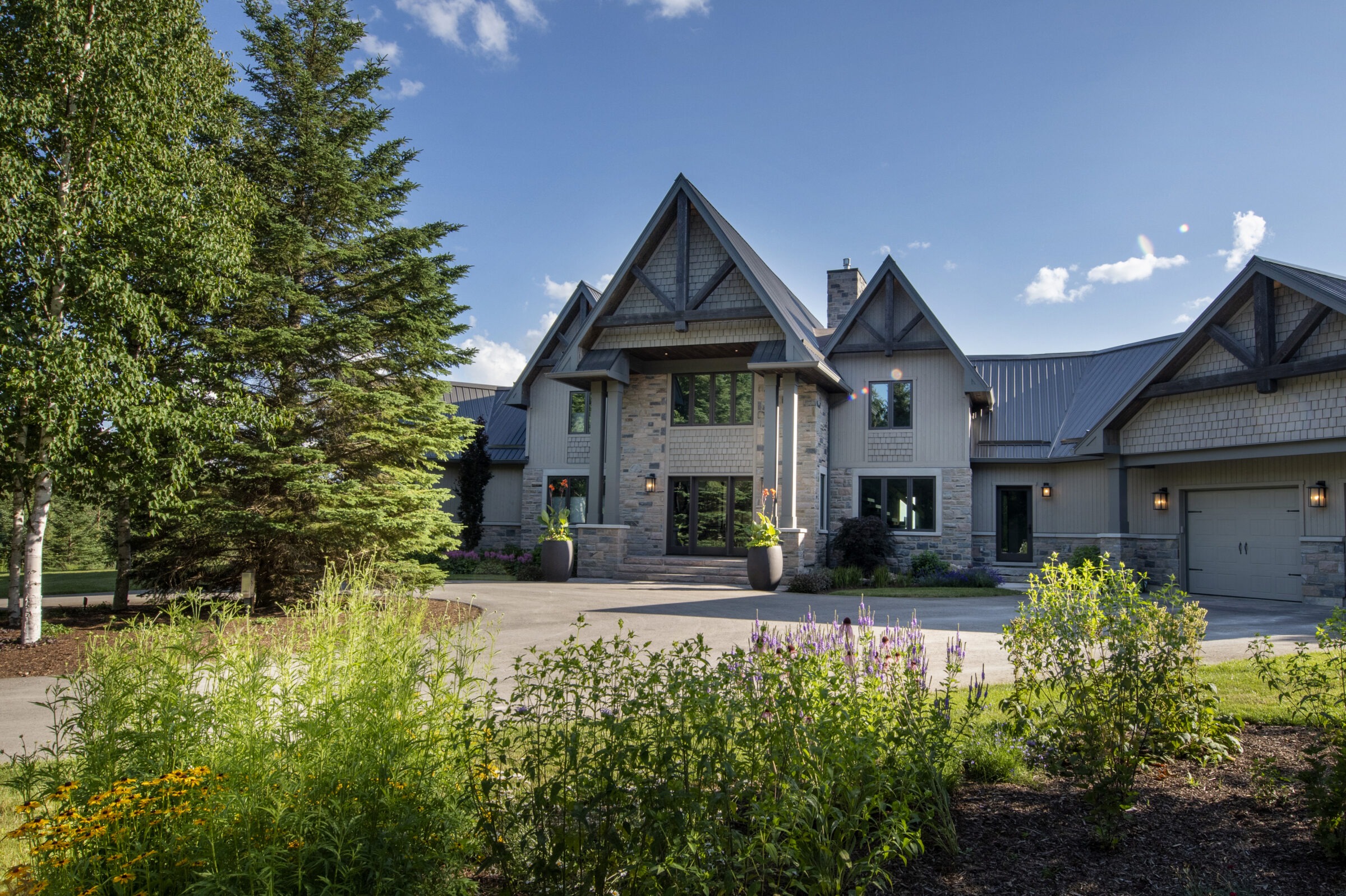 A modern house with a stone facade and gabled roof is surrounded by lush garden greenery under a clear blue sky.