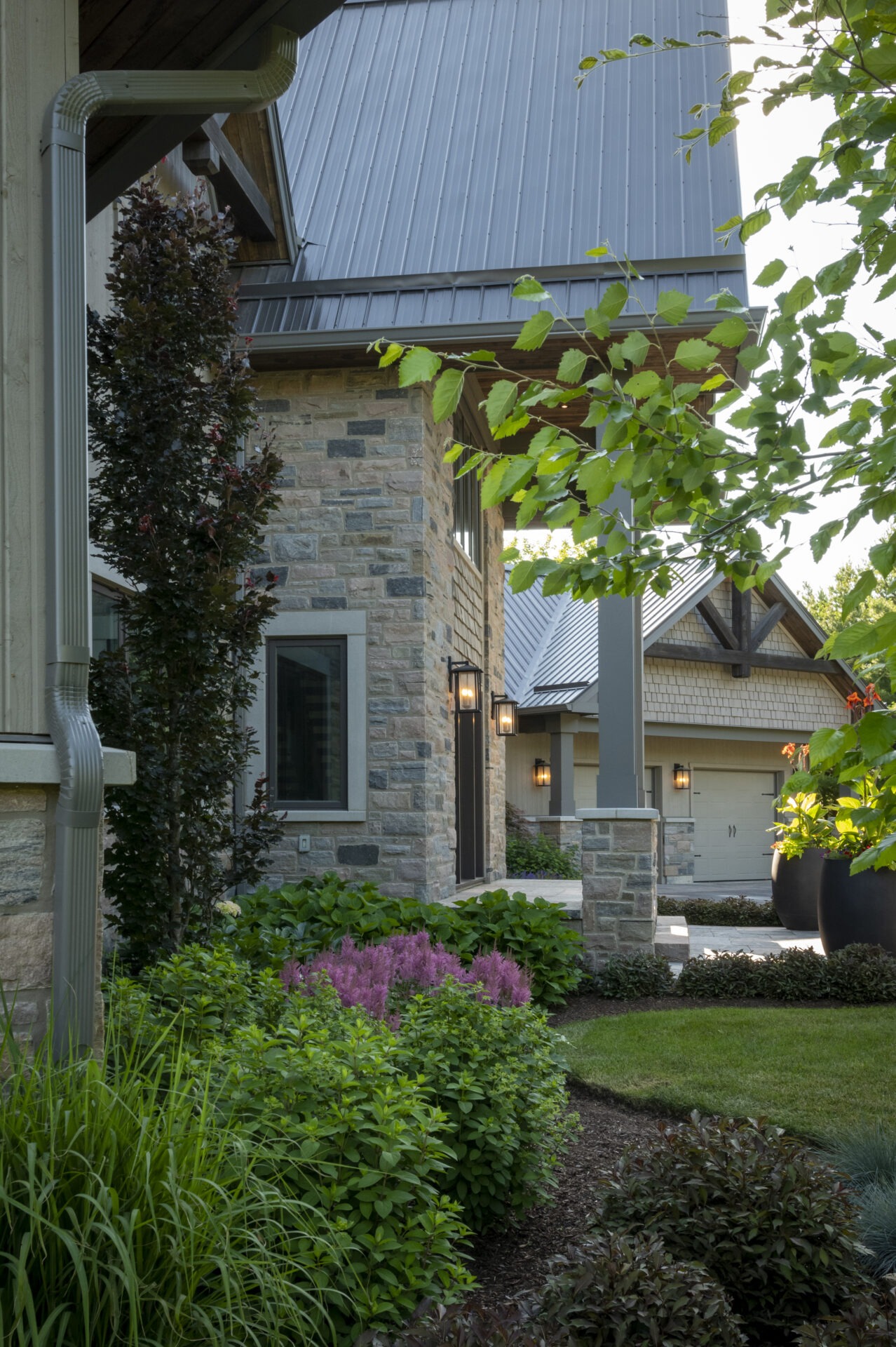 Modern house with stone facade, metal roof, and vibrant landscaped garden with lush greenery and colorful flowers. Quiet, residential setting.