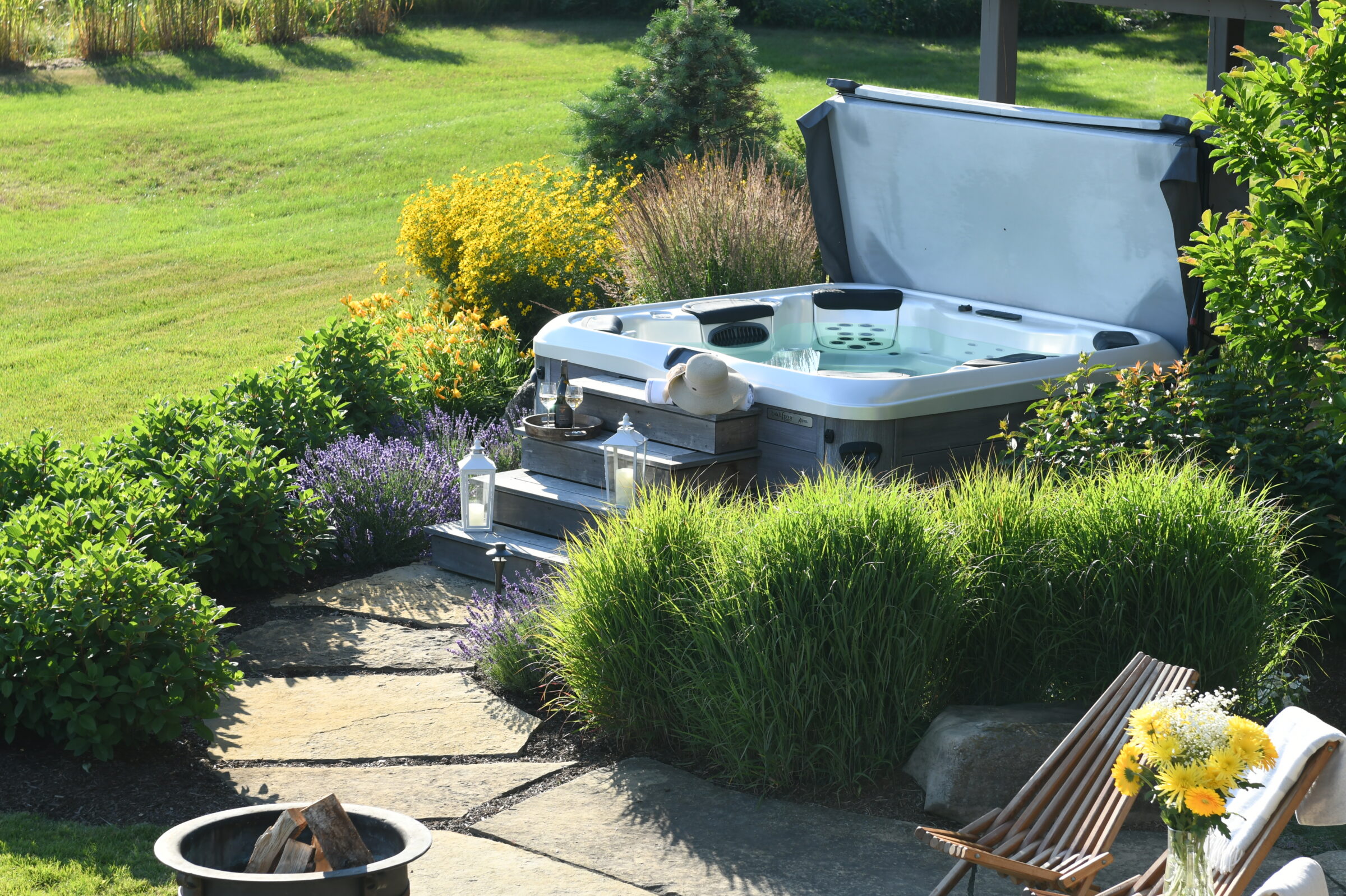 A hot tub in a garden setting with lush greenery, colorful flowers, and a cozy seating area with a fire pit nearby.