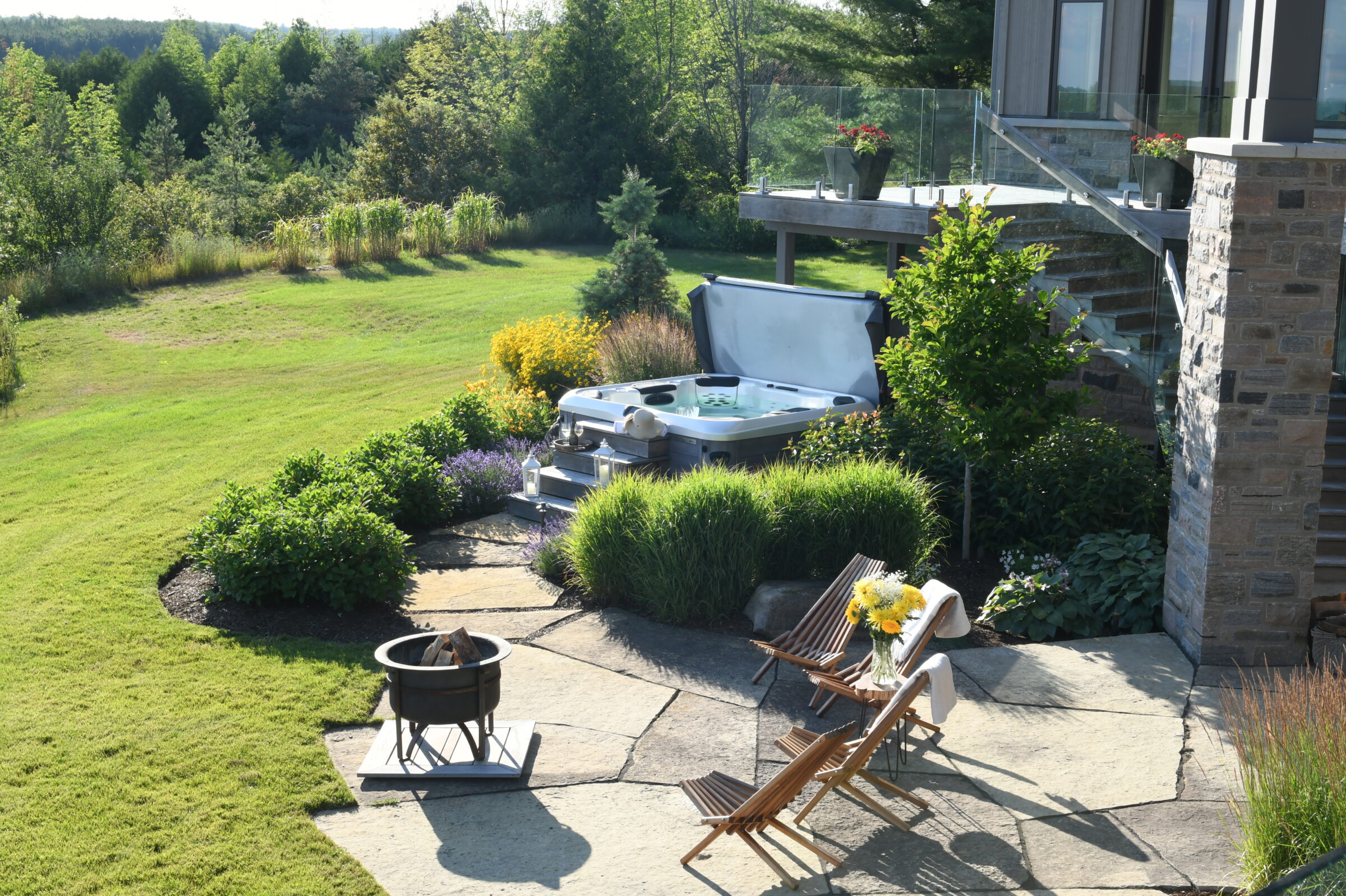 Spacious backyard with vibrant greenery, a hot tub, two wooden chairs, and a small fire pit on a stone patio.