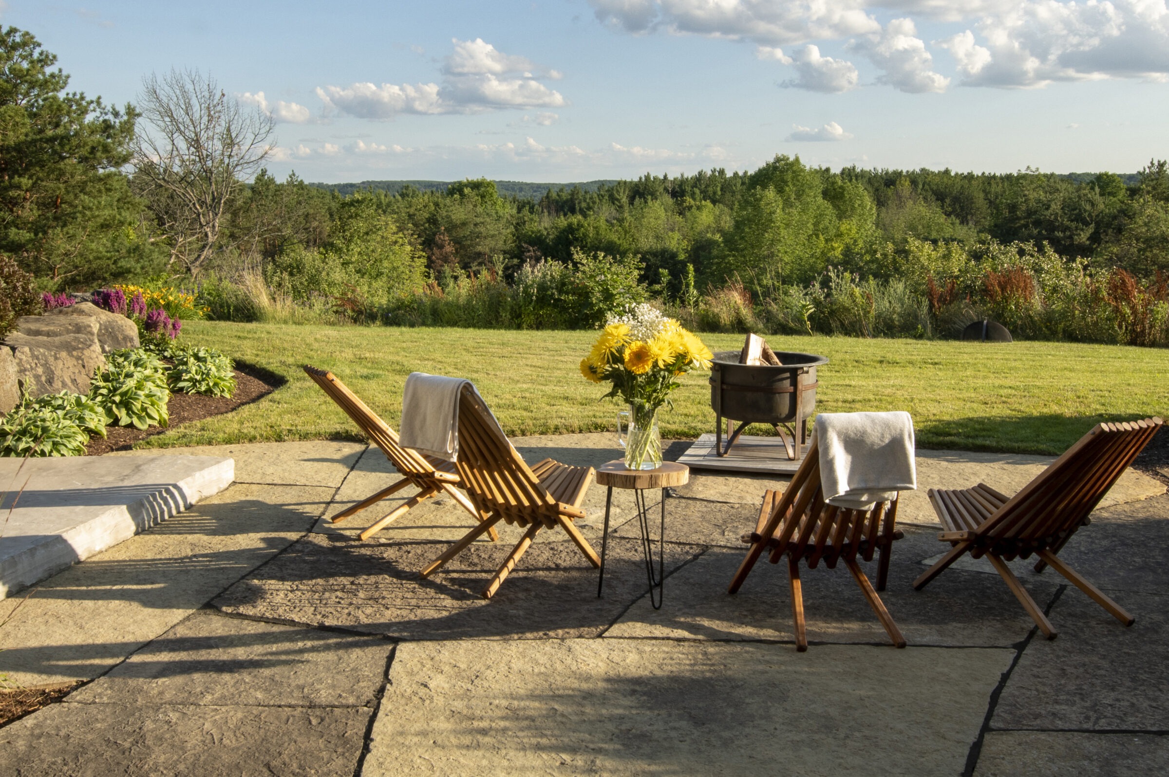 A scenic patio with wooden chairs, table, and sunflowers in a vase overlooks a lush green landscape under a partly cloudy sky.