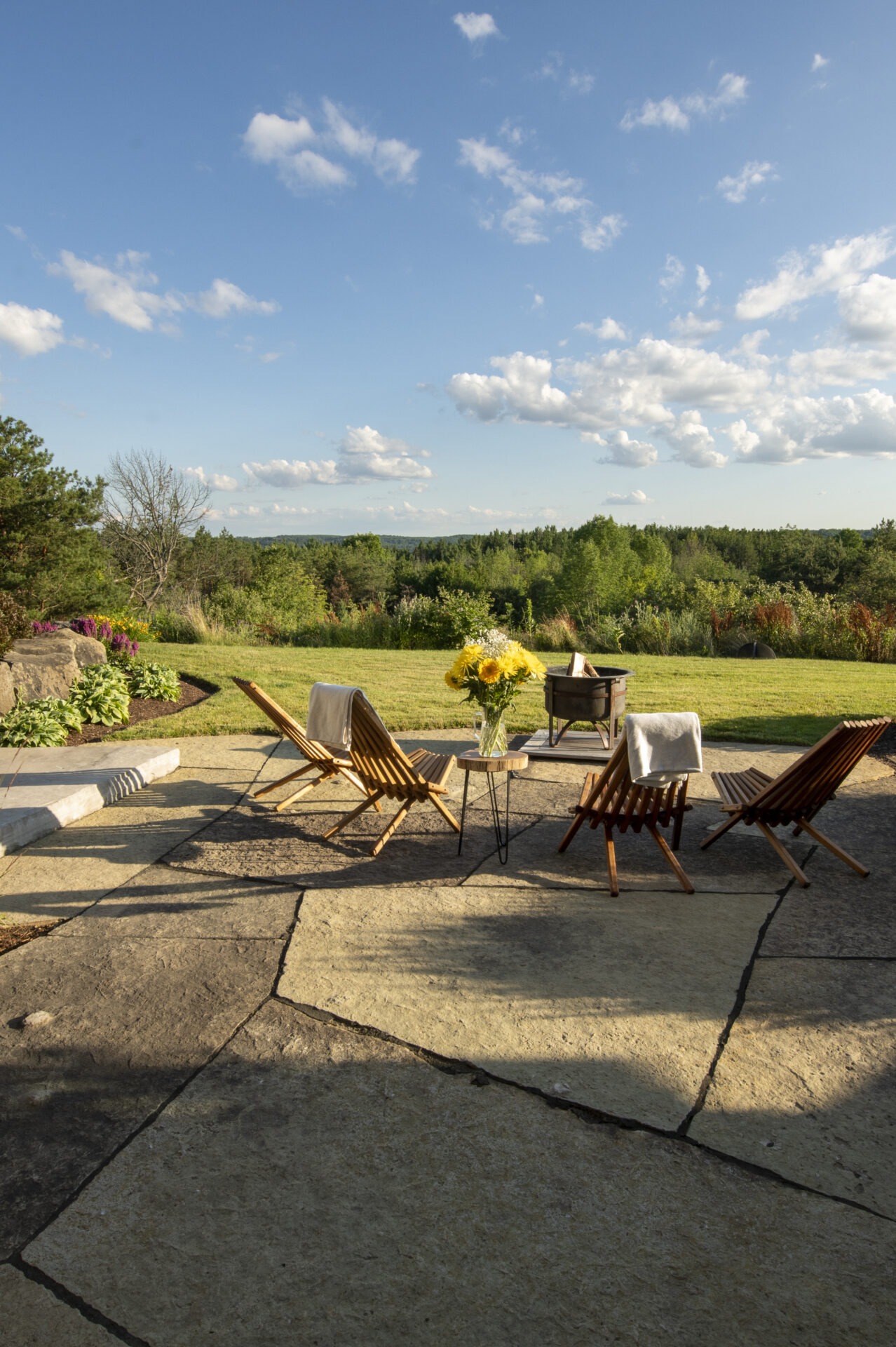 A serene patio scene with wooden chairs, sunflowers, and a fire pit, overlooking a lush, green landscape under a bright, blue sky.