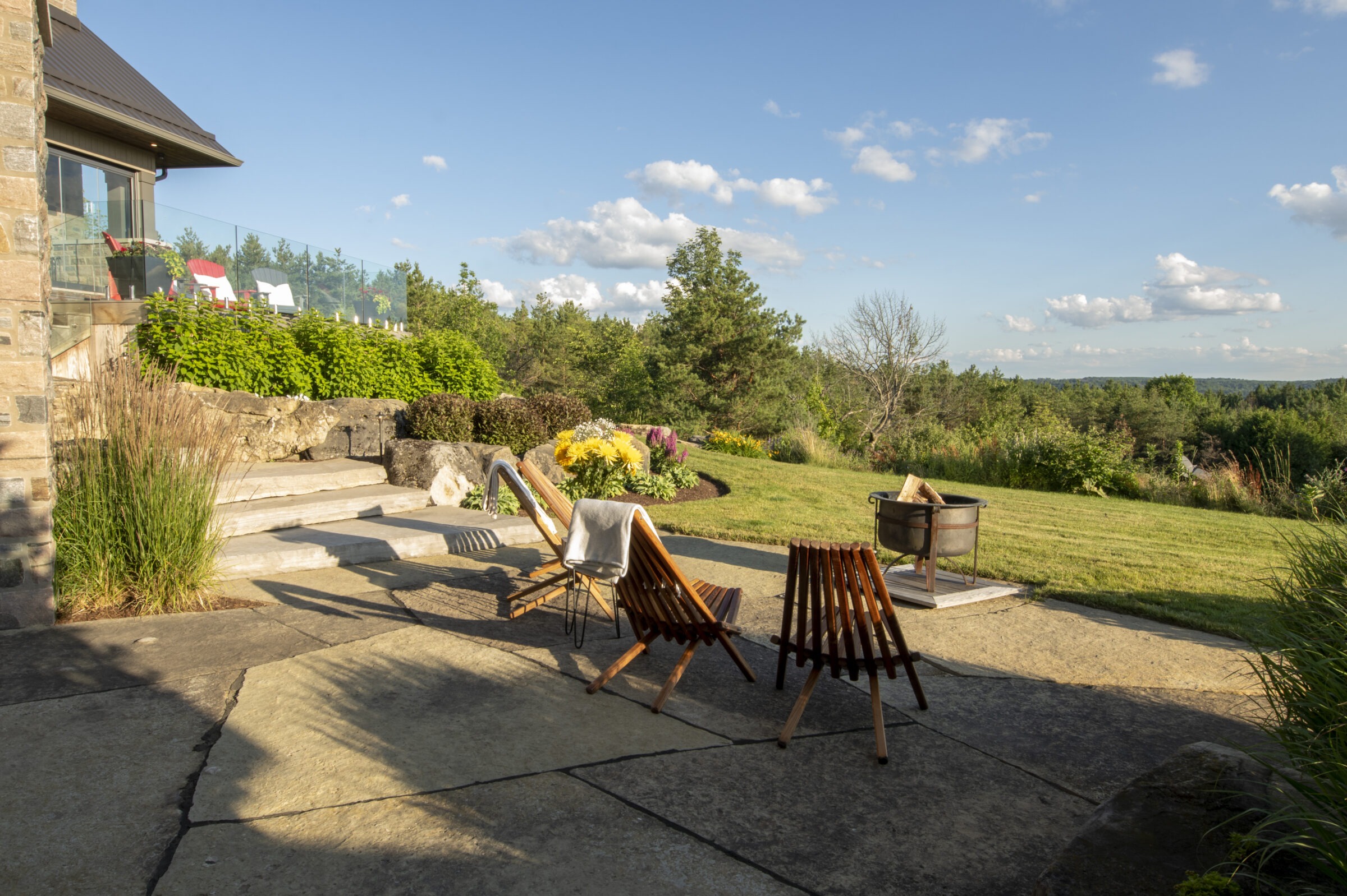 A scenic patio setting with wooden chairs, a fire pit, and lush greenery under a clear blue sky. Luxurious outdoor relaxation spot.