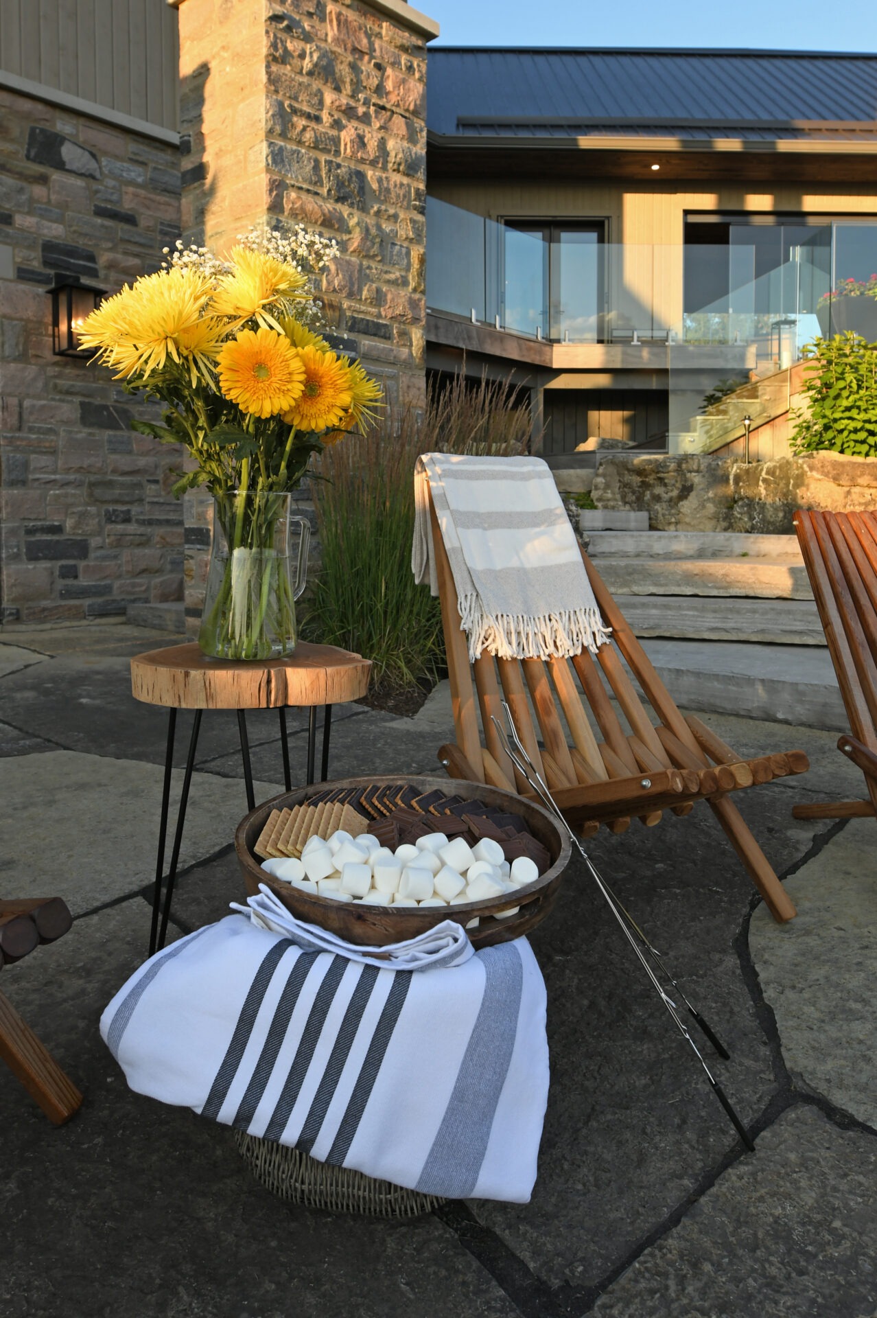 A cozy outdoor setting features wooden chairs, a table with flowers, and a bowl of s'mores ingredients, set against a stone building.