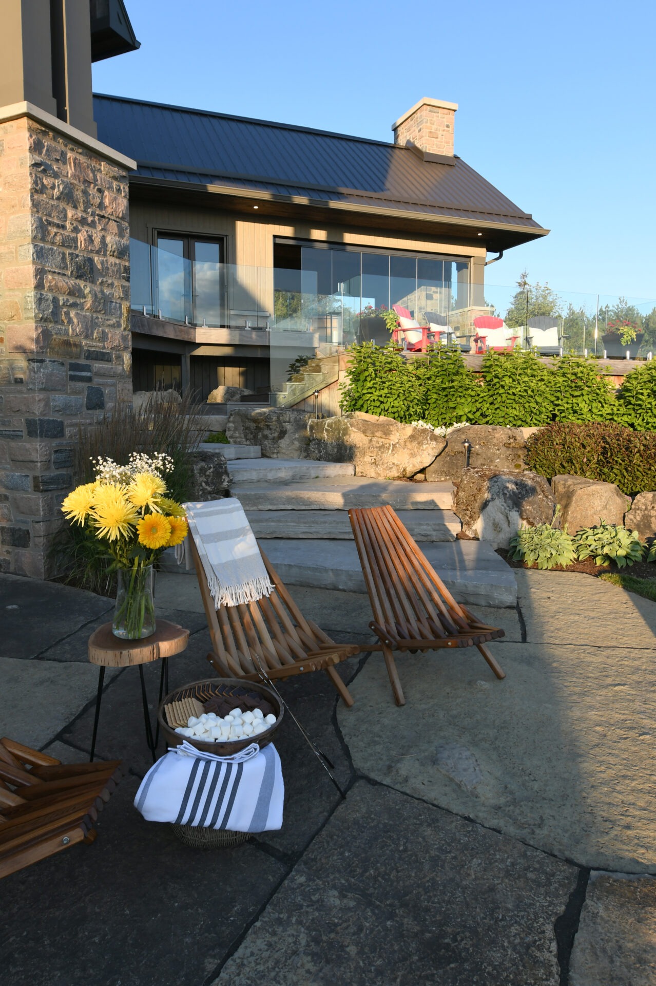 Modern patio with wooden chairs, stone steps, and flowers. A glass railing enhances the contemporary house with lush greenery. Relaxed outdoor setting.