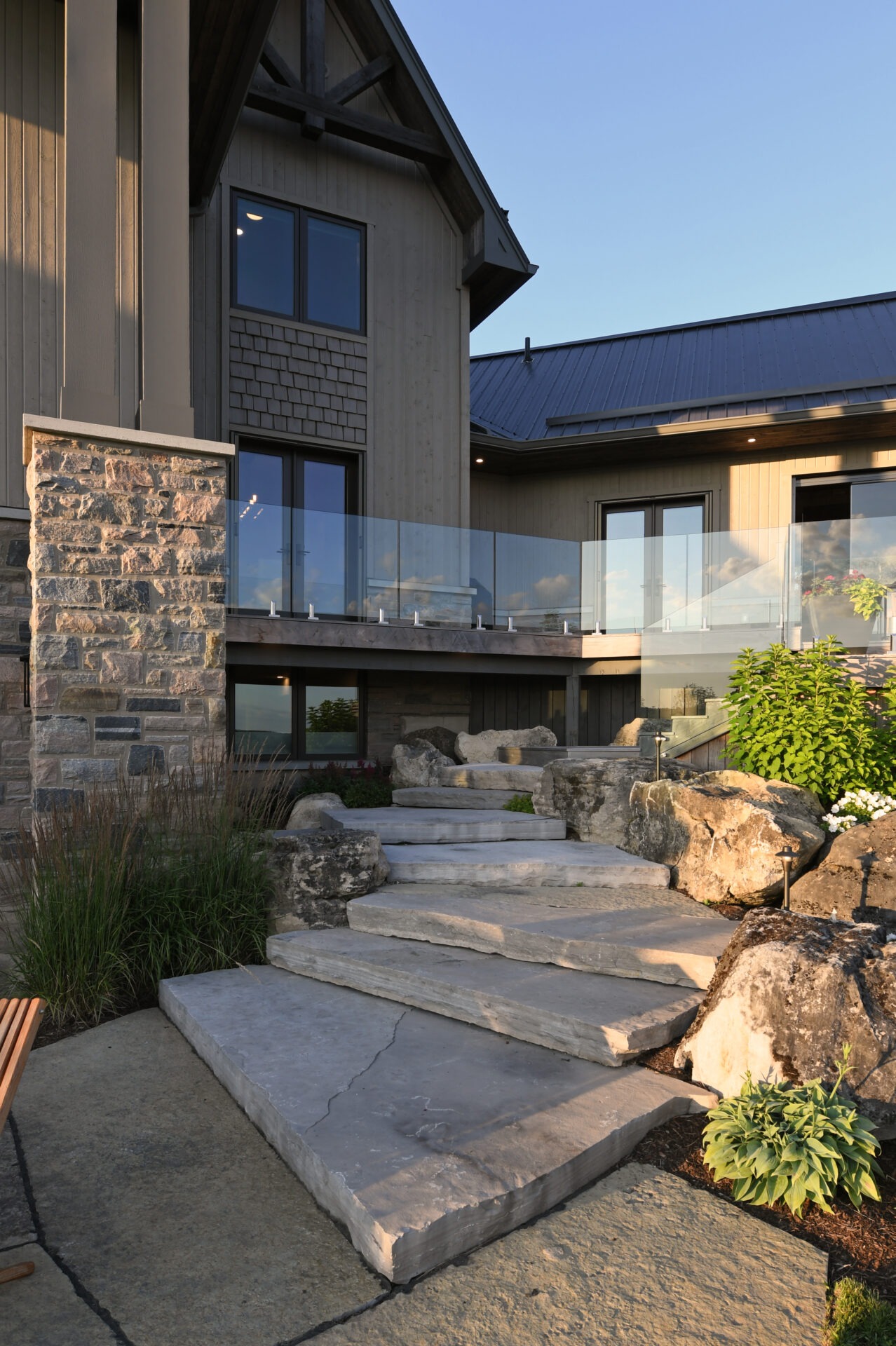 Modern house exterior with stone steps, glass railing, and landscaped garden. The building features a wooden facade and large windows, bathed in sunset light.