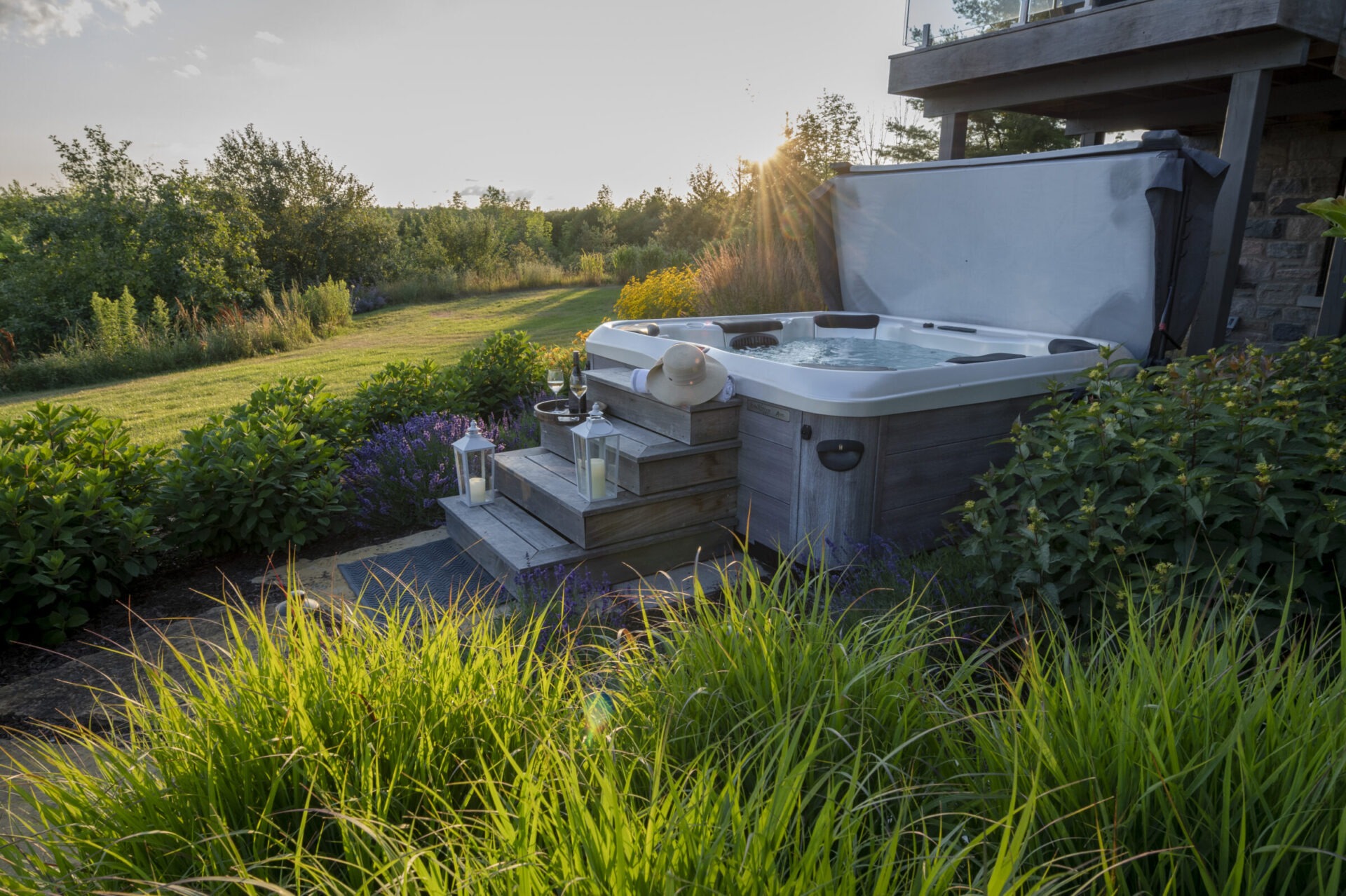 A hot tub sits on a wooden deck surrounded by lush greenery and candles, with sunlight filtering through the trees in the background.