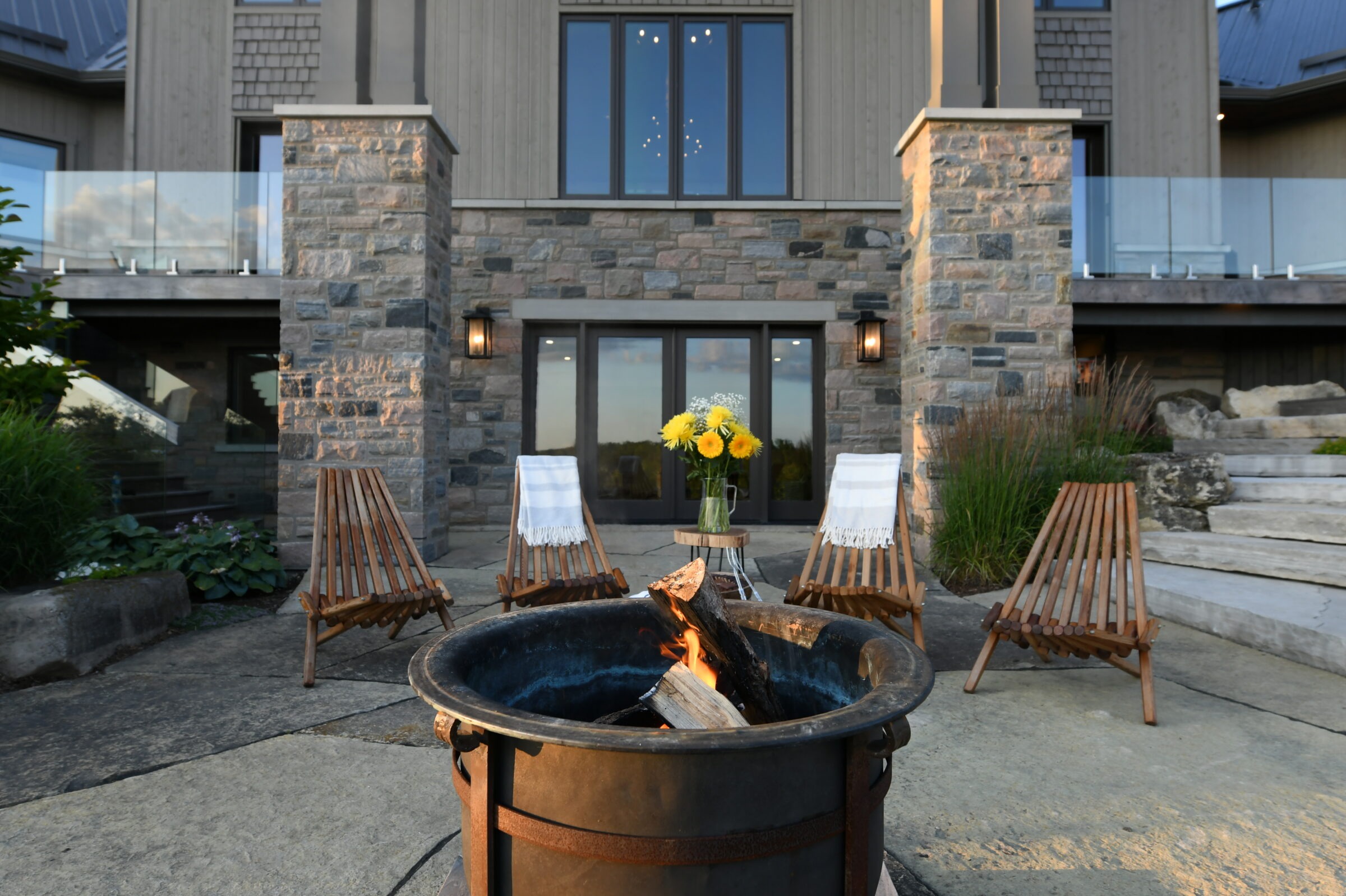Stone patio with fire pit, four wooden chairs, and sunflowers in vase. Large glass door entry of modern home in background.
