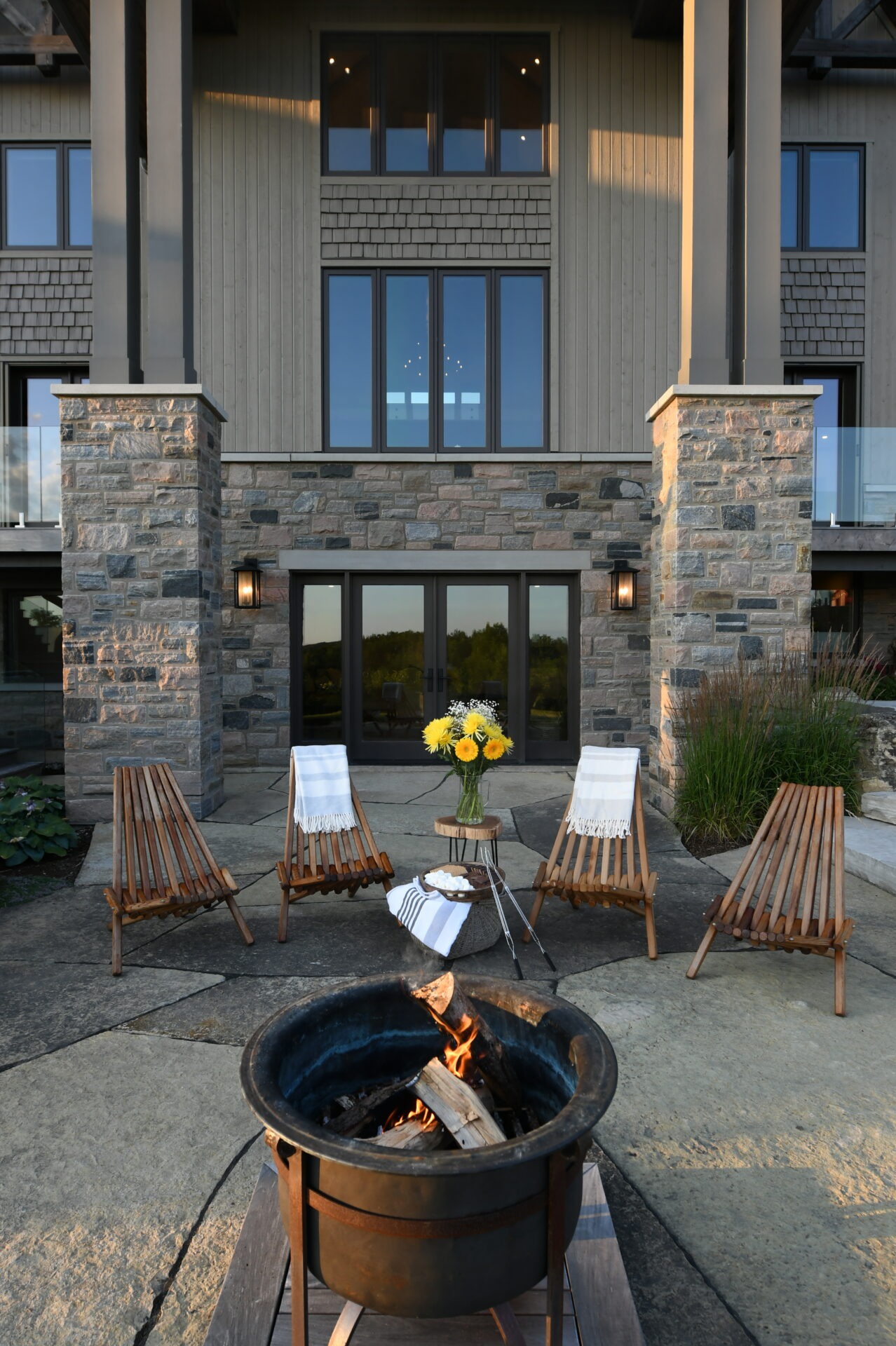 Cozy patio with a fire pit, wooden lounge chairs, sunflowers on a side table, and a stone building facade in the background.