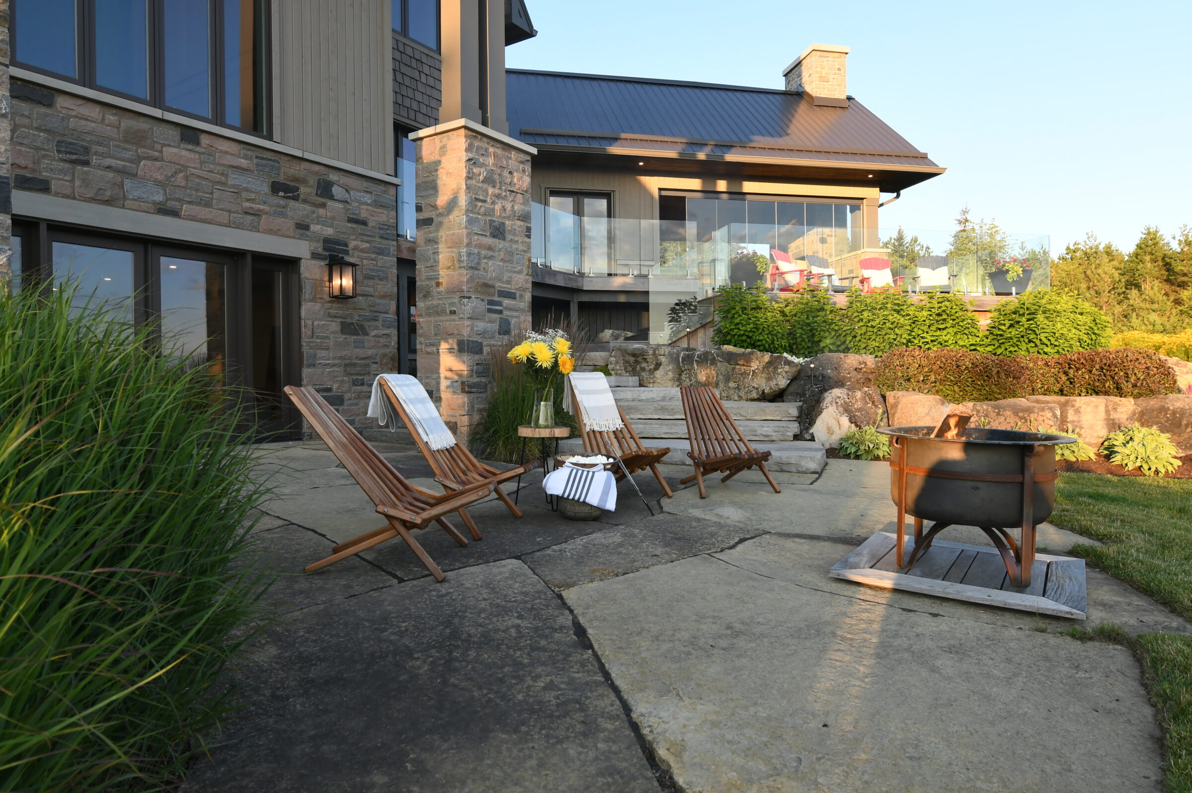 Modern house with stone patio features wooden chairs, a fire pit, and vibrant landscaping. Sunlight casts soft shadows on the scene.