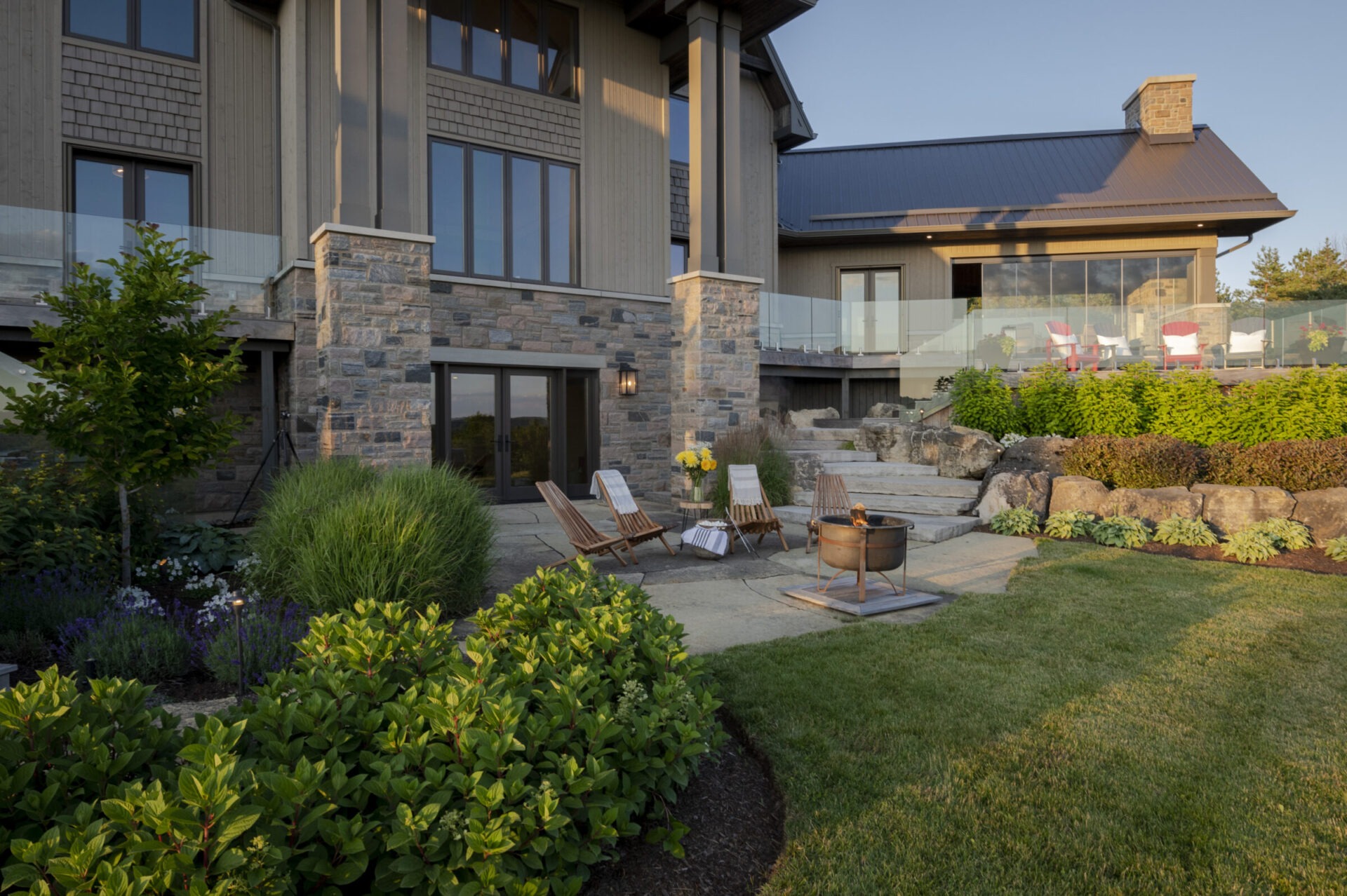 Modern house with a stone facade, glass railings, patio chairs, fire pit, surrounded by lush greenery and well-manicured garden.
