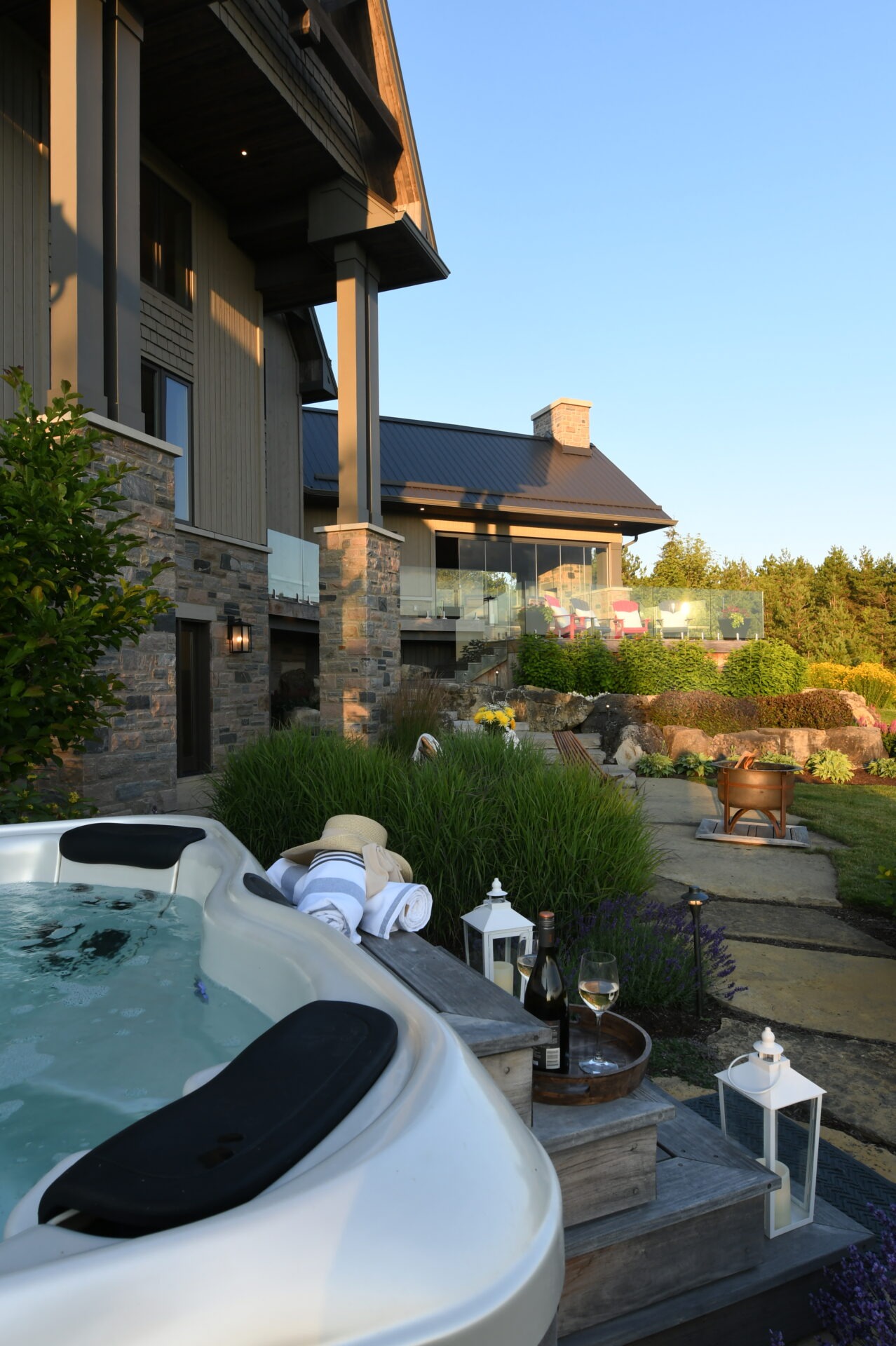 A modern house with stone facade, hot tub, wine, and towels. Relaxing outdoor setting with lush greenery and clear blue sky.