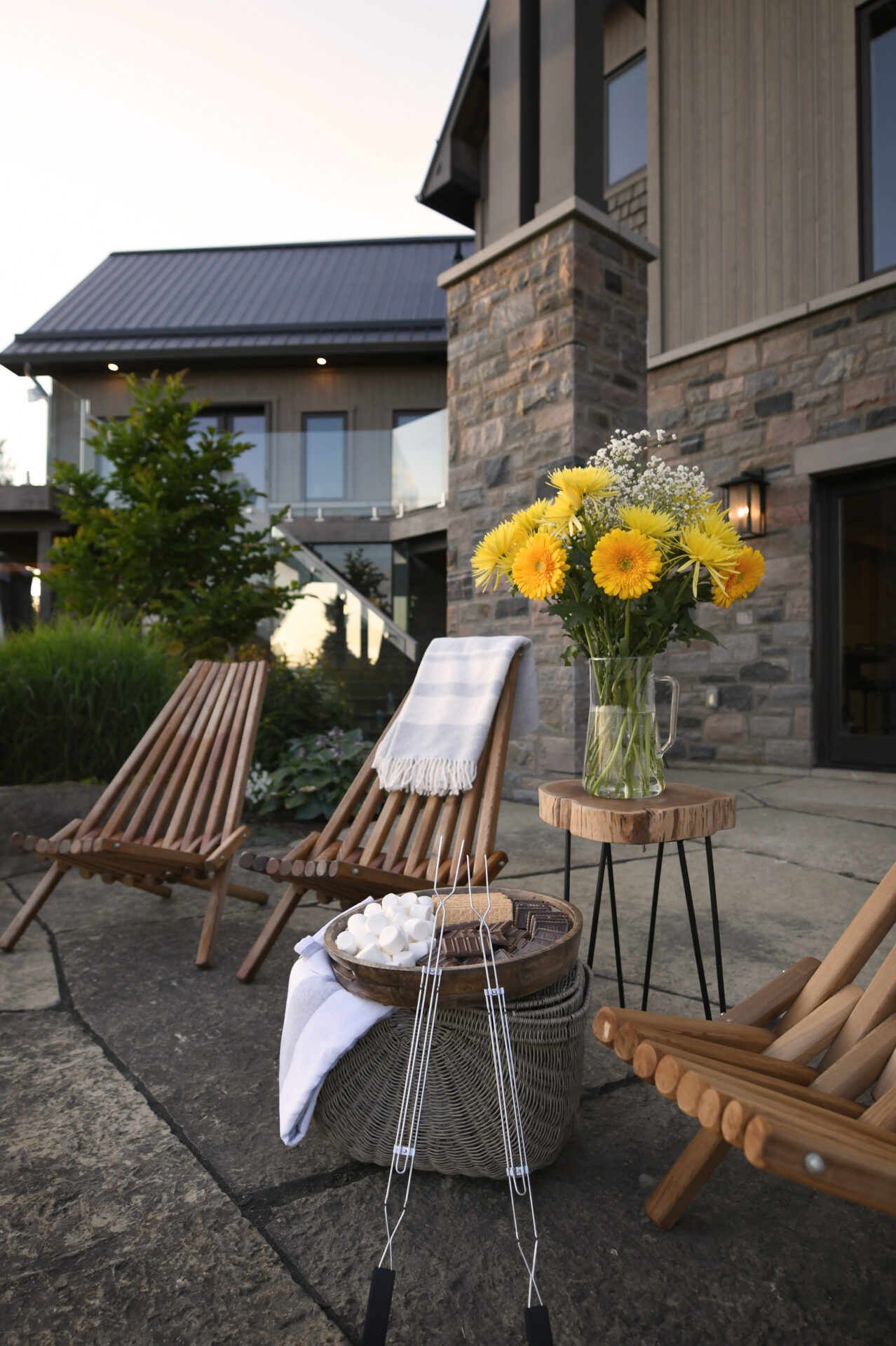 Wooden chairs and a table with flowers and marshmallows set on a stone patio outside a modern house with a glass balcony.