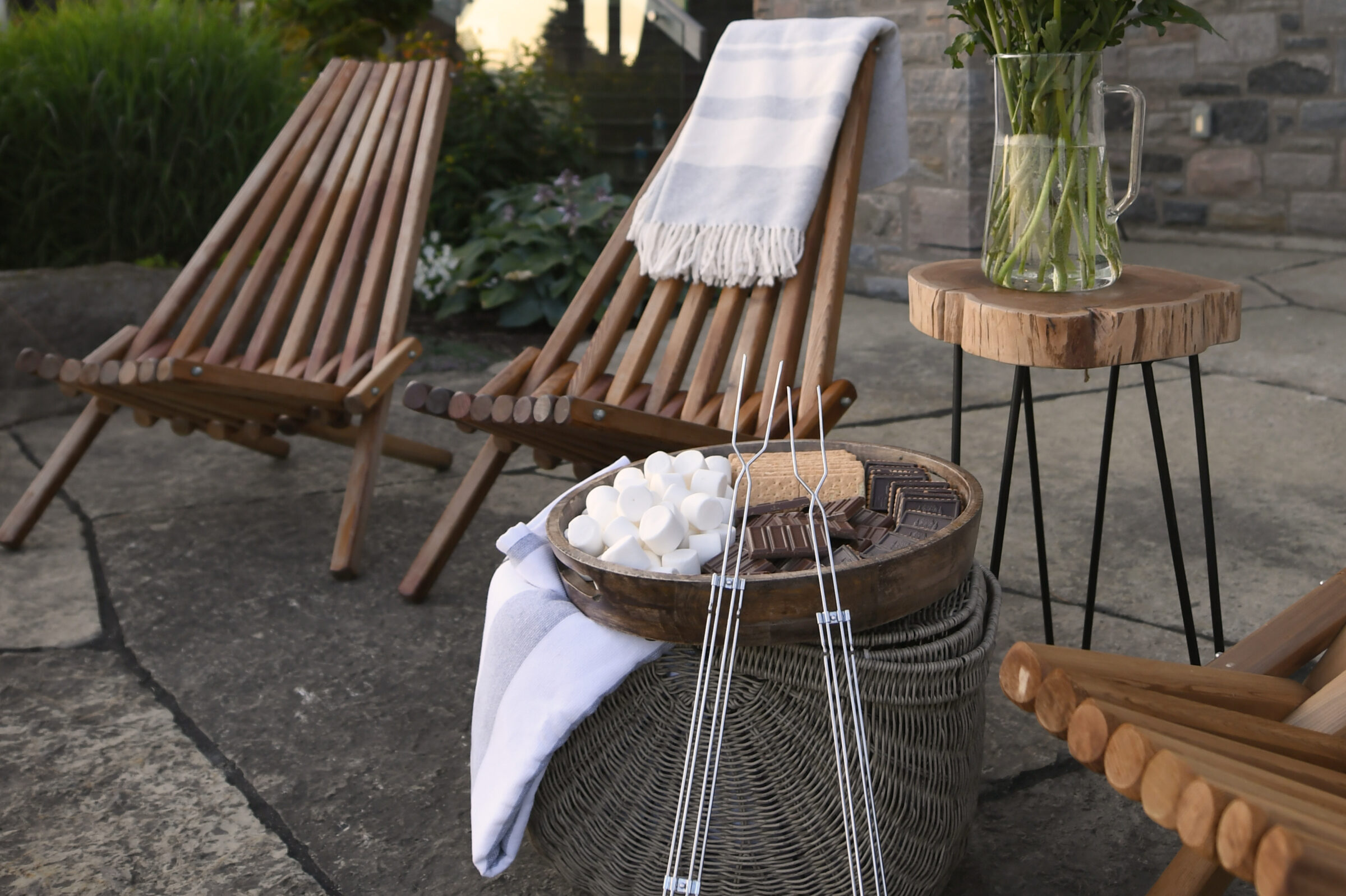 Wooden chairs and side table on patio, basket with marshmallows, chocolate, and skewers, striped blanket draped over chair; relaxing outdoor setup.
