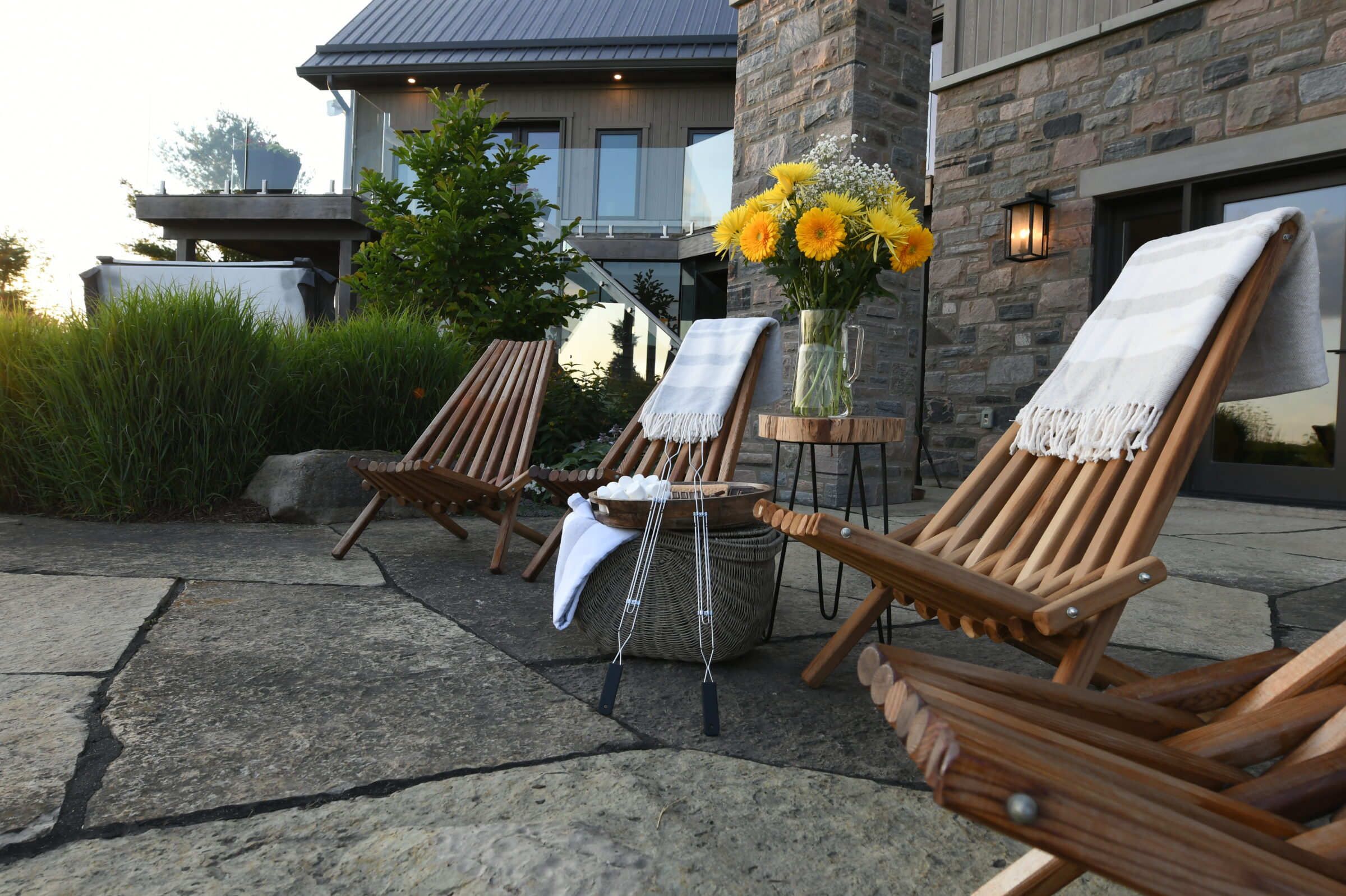 Patio with three wooden lounge chairs, a side table with yellow flowers, and a stone building facade in the background. Peaceful outdoor setting.