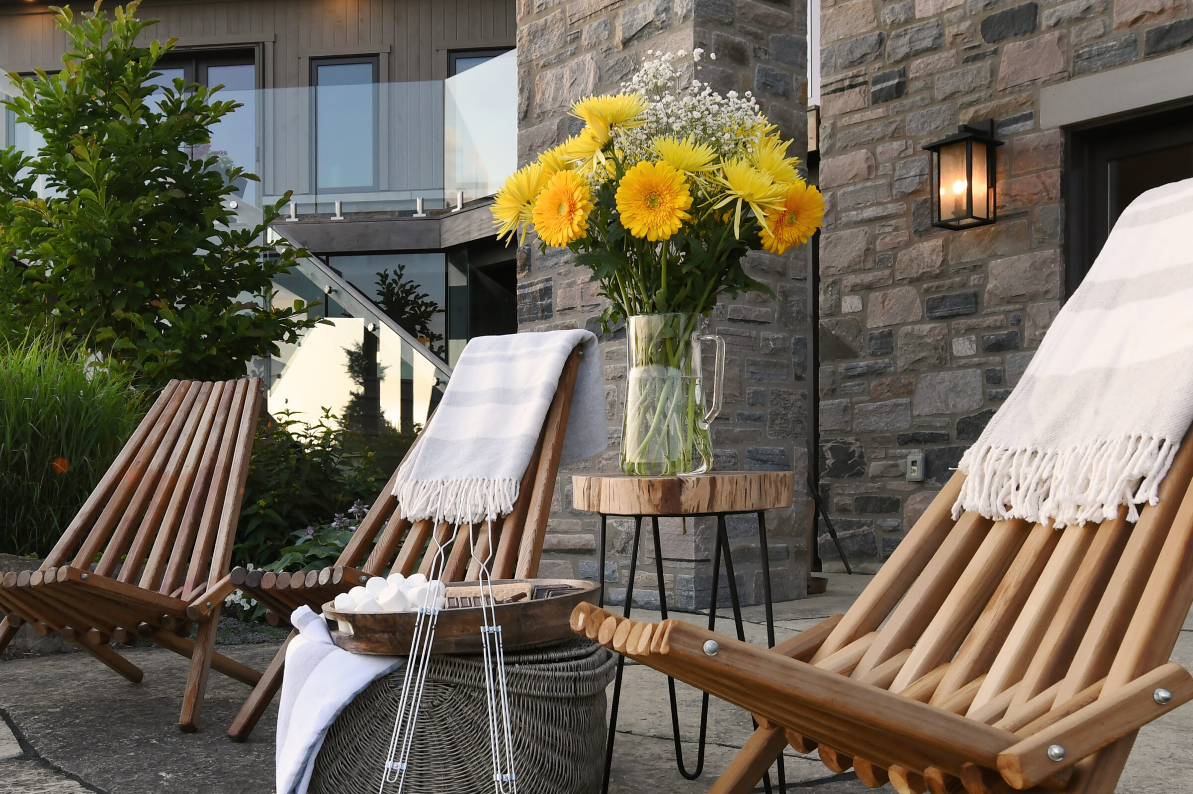 Two wooden chairs, a side table with yellow flowers, and blankets create a cozy outdoor setting by a stone wall and glass railing.