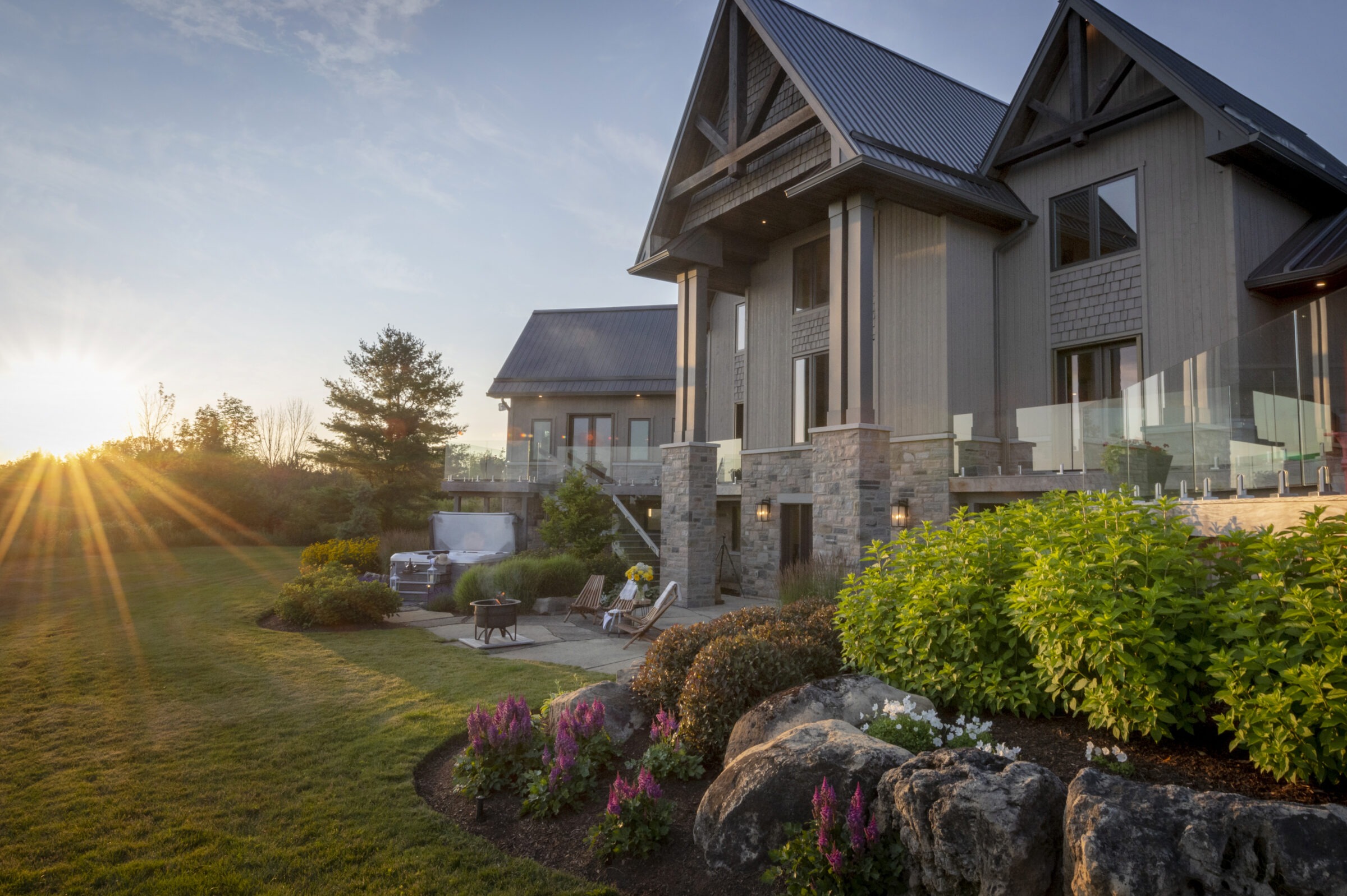 Modern house with stone and wood facade, lush garden, patio furniture, and vibrant flowers. Sun setting behind trees, creating warm, golden light.