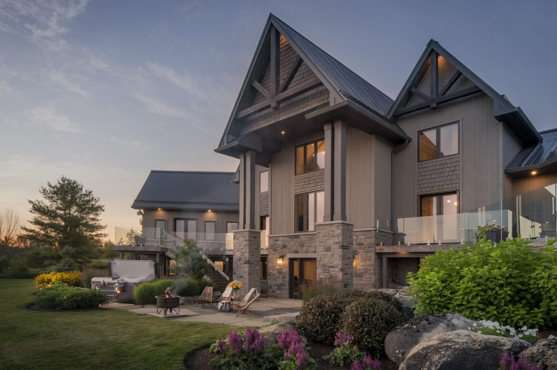 Modern house with stone and wood exterior, large windows, and a landscaped yard at sunset. Outdoor seating and fire pit visible.