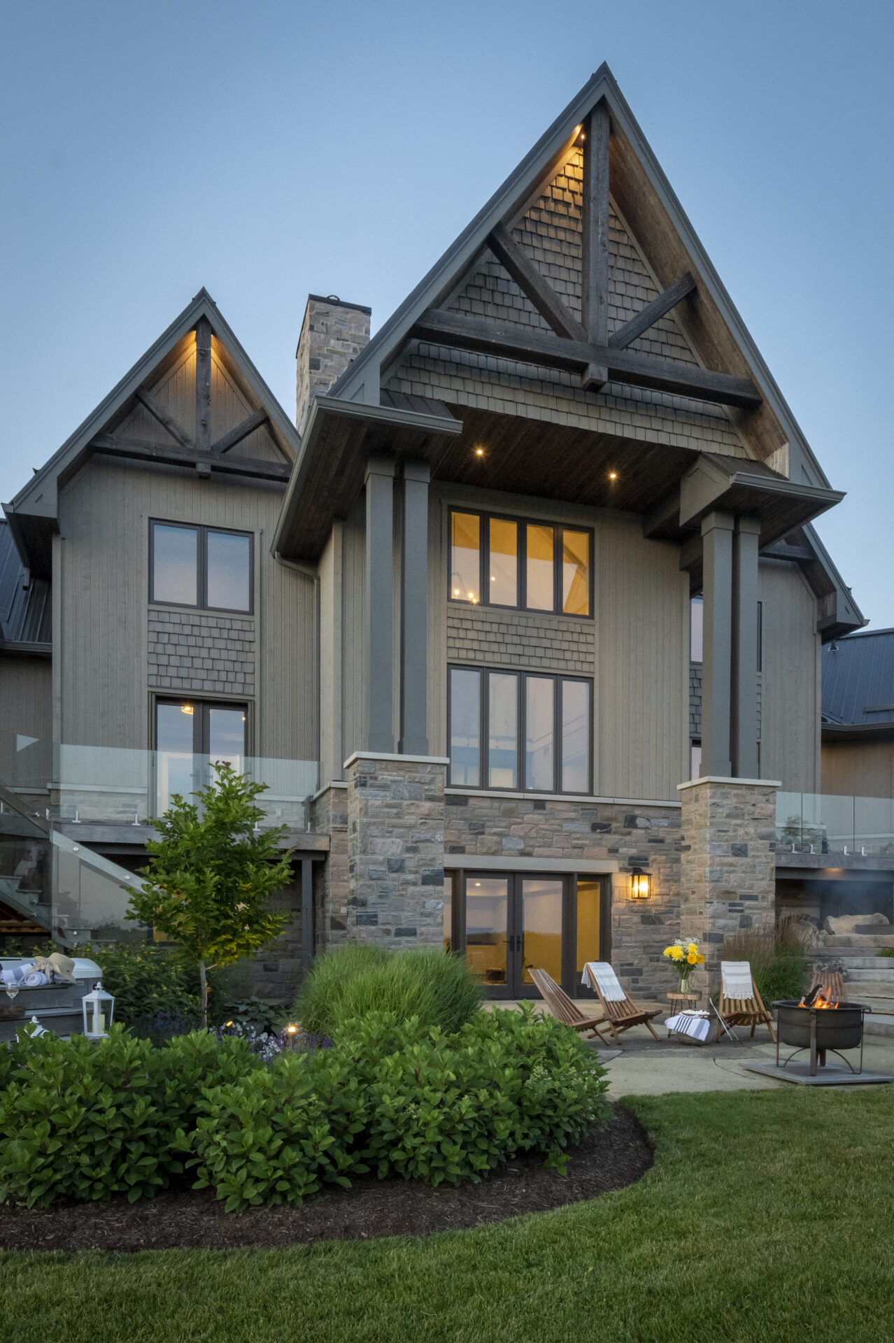 Large two-story rustic house with stone facade, wooden details, and triangular roofs. Lush garden and cozy patio with chairs and firepit.