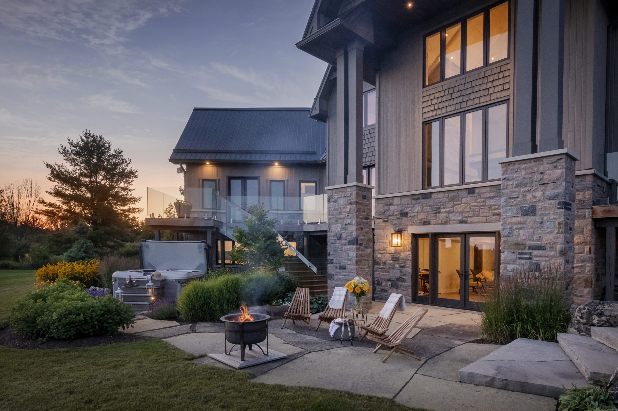 Modern house at sunset with stone facade, outdoor seating around a fire pit, and hot tub surrounded by lush garden and open sky.