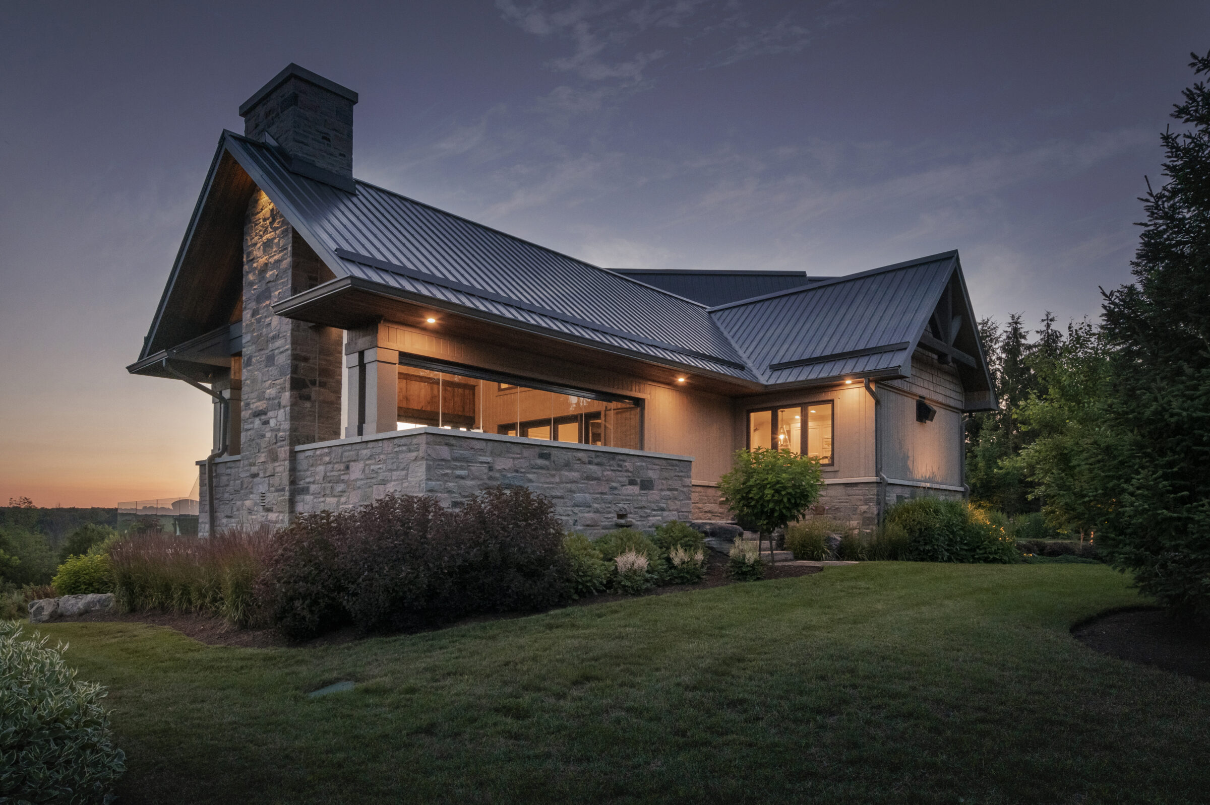 Modern house with stone and wood exterior, sloped metal roof, surrounded by lush greenery. Evening sky provides a serene backdrop.