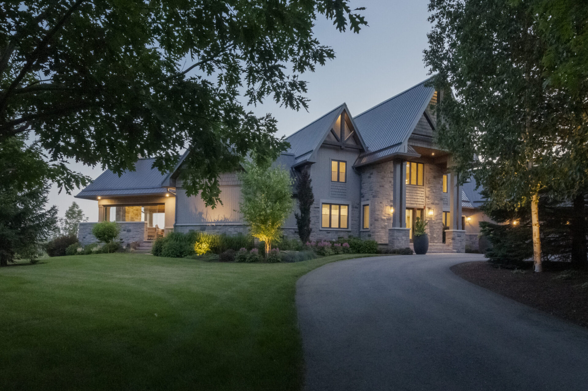 A large, elegant house with a modern design is illuminated in the evening, surrounded by lush greenery and a curved driveway.