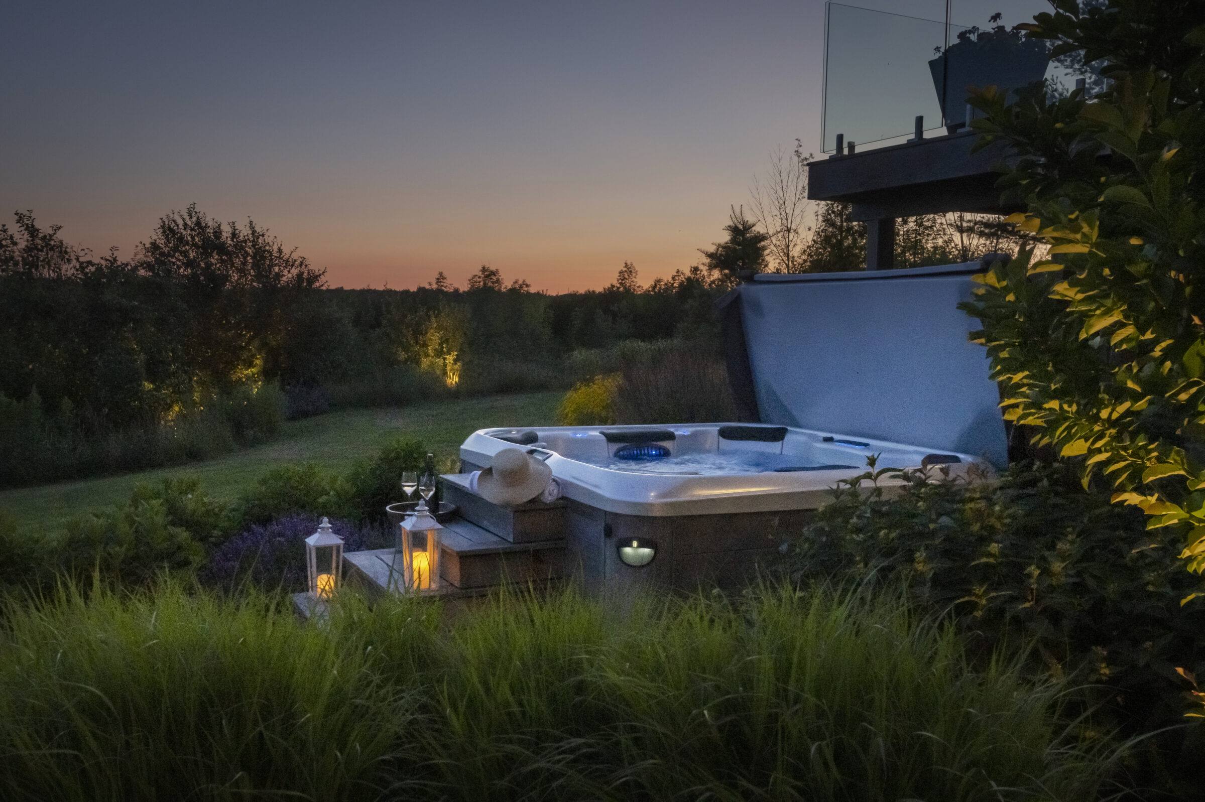 Hot tub on a deck at dusk, surrounded by glowing lanterns and greenery. A serene landscape with trees under a colorful evening sky.