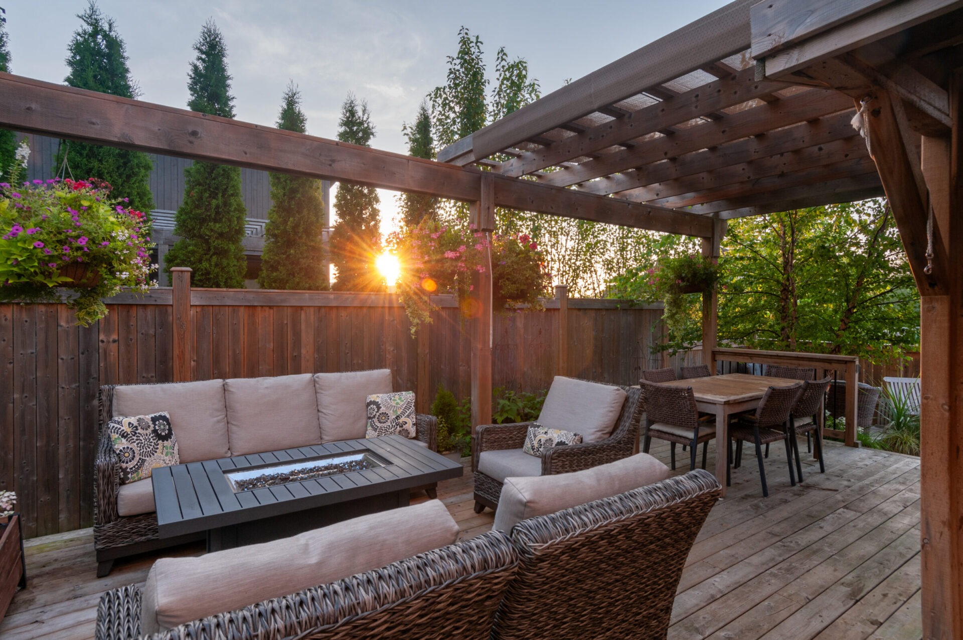 Outdoor patio with wicker furniture, wooden pergola, and dining table. Sun setting over trees, casting warm light on the cozy space.