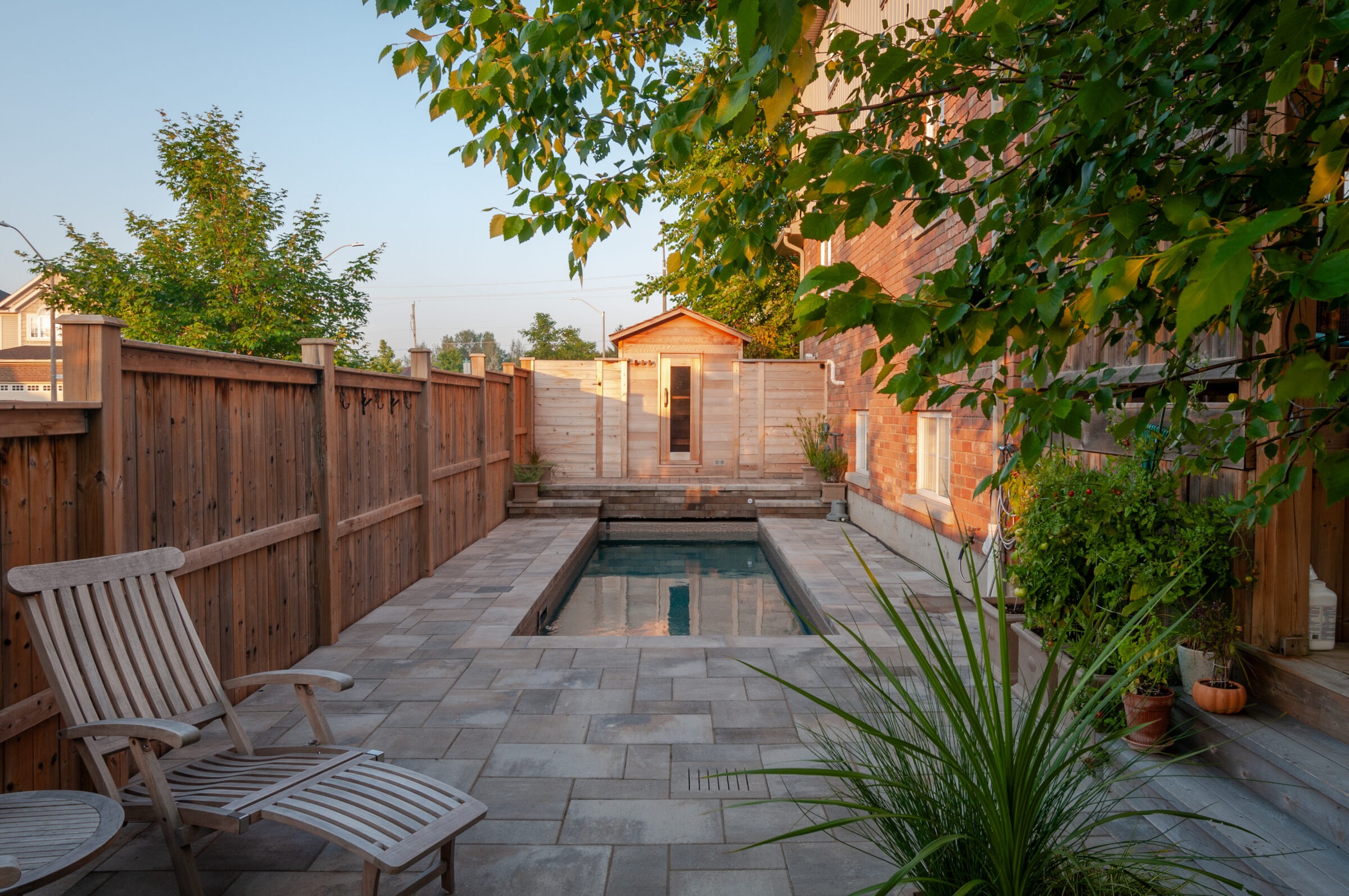 A serene backyard features a small pool, wooden fence, and lush plants, with a wooden sauna structure at the far end.