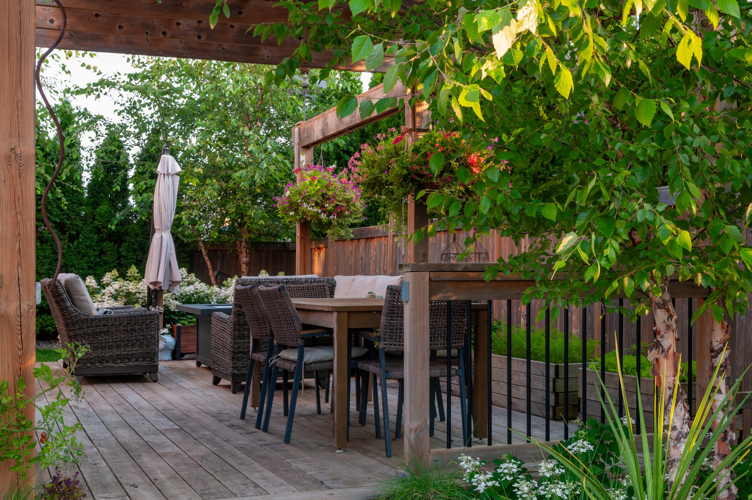A cozy wooden deck with wicker furniture, under a pergola surrounded by lush greenery and vibrant flowers, creating a tranquil outdoor space.