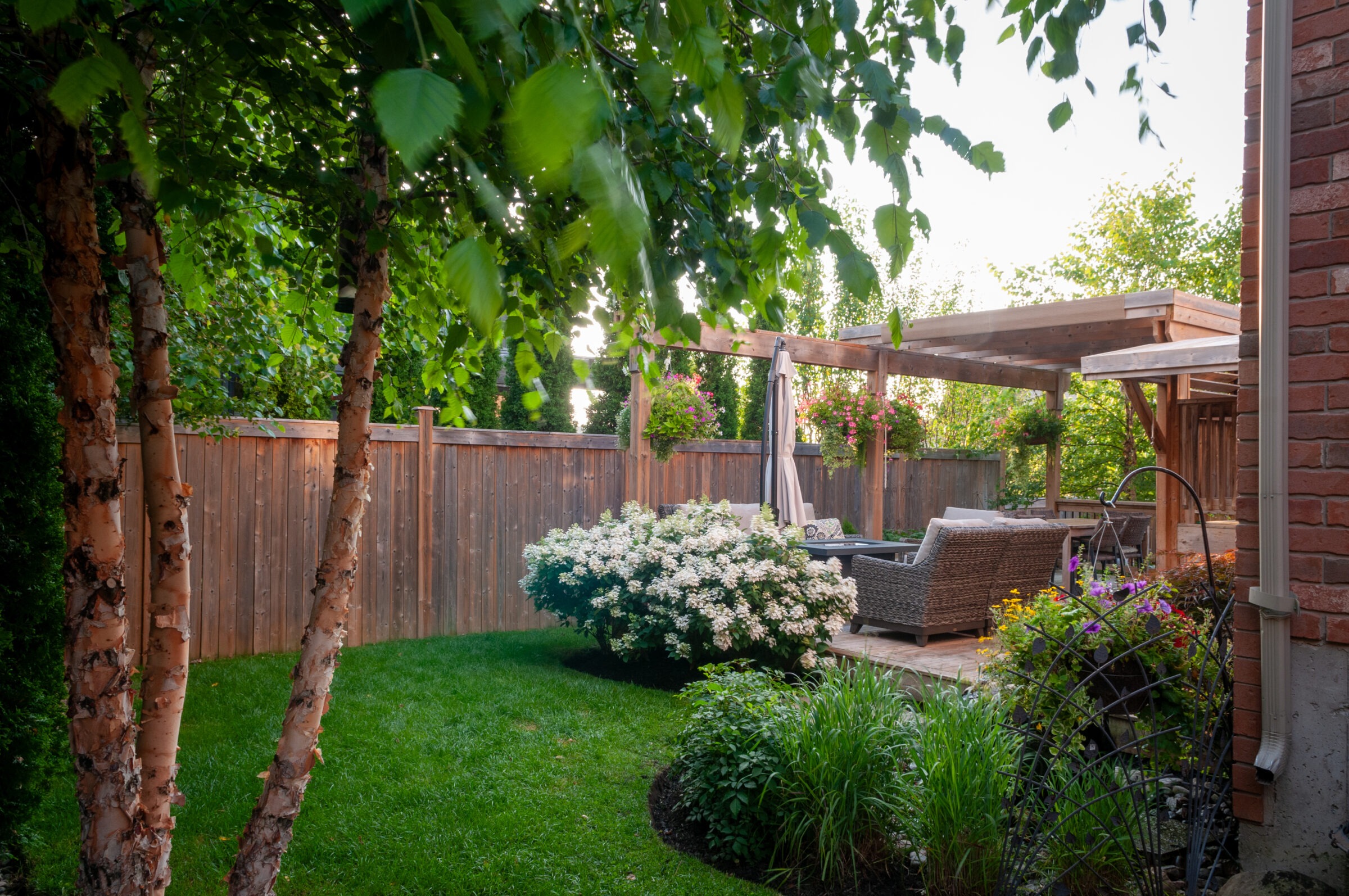 A lush backyard garden with trees, flowers, and a cozy seating area under a wooden pergola, fenced by a wooden barrier.
