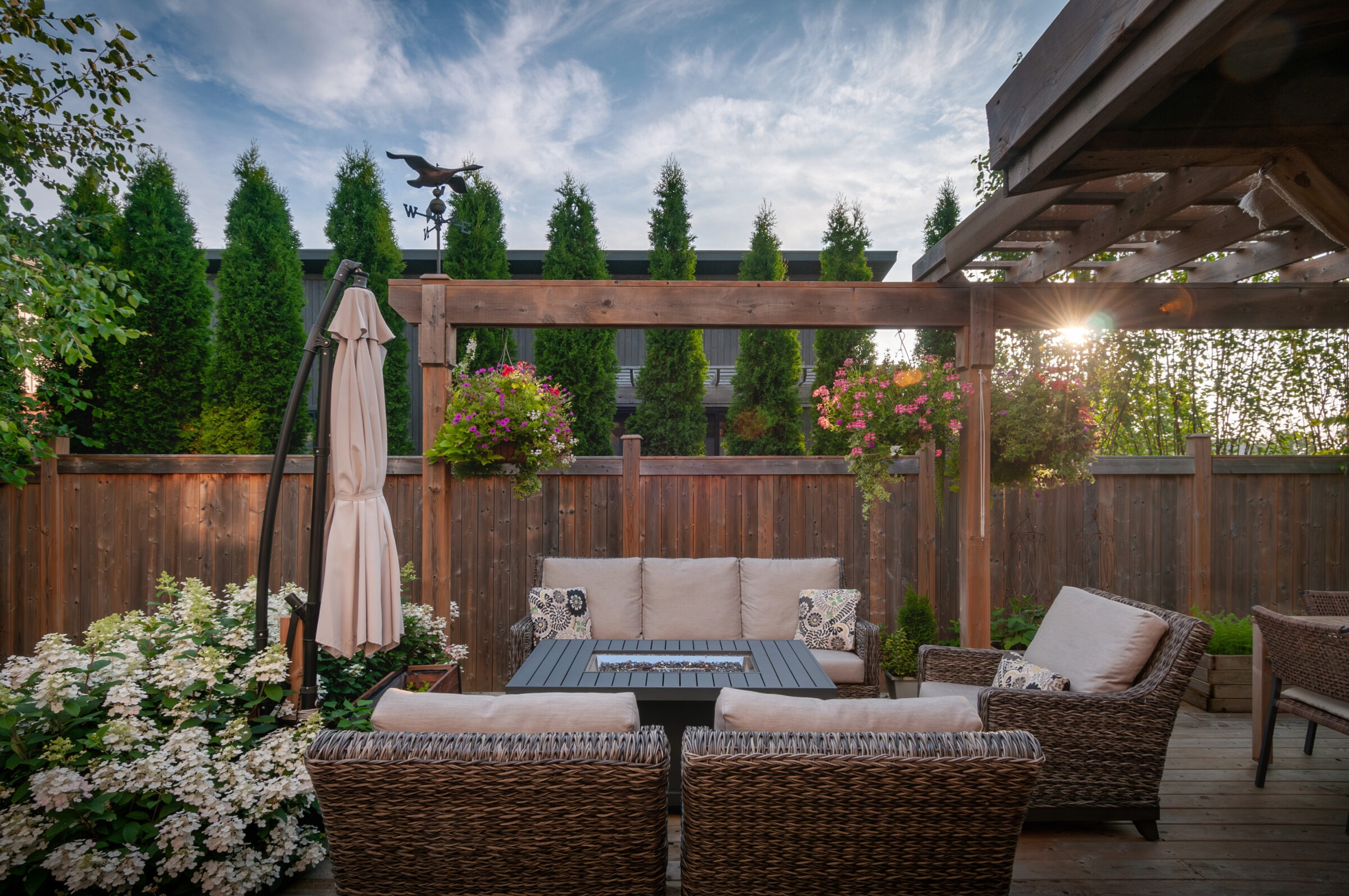 Outdoor patio featuring wicker furniture, a pergola, and lush greenery. Hanging flower baskets add color; weather vane tops the scene under blue sky.