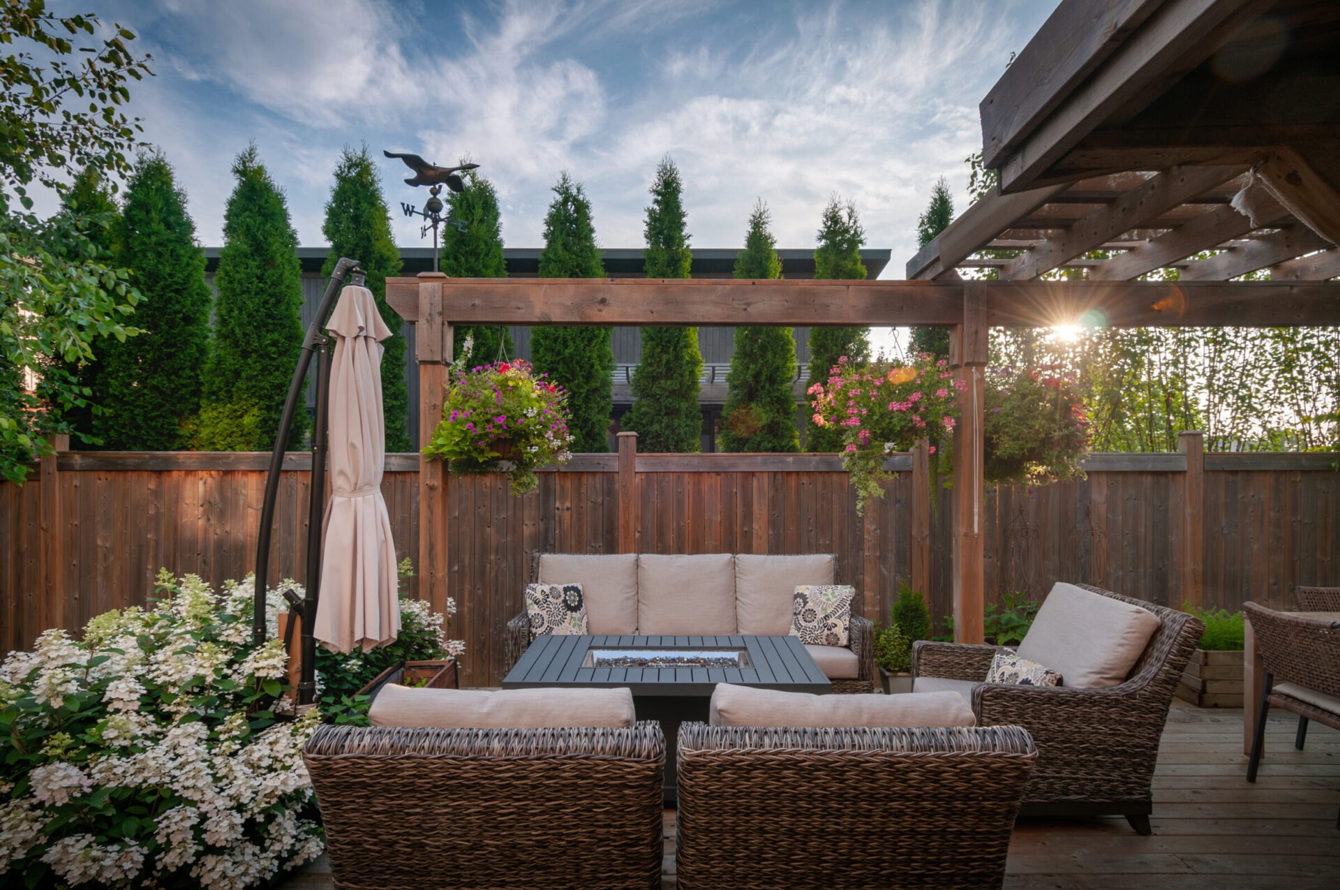 Cozy patio with wicker furniture, flowers, and a pergola. Surrounded by trees and a high wooden fence. Clear sky and a weather vane.