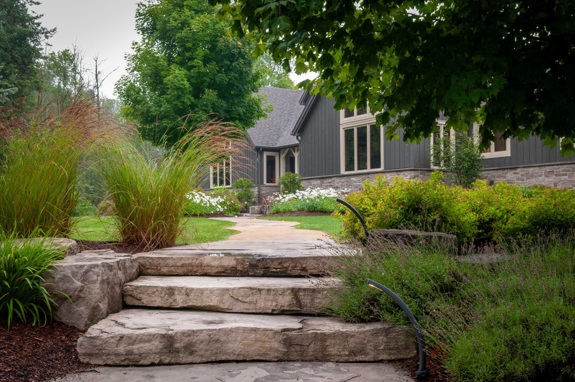A rustic stone walkway leads to a stylish home surrounded by lush greenery and flowering plants, creating a serene, inviting atmosphere.