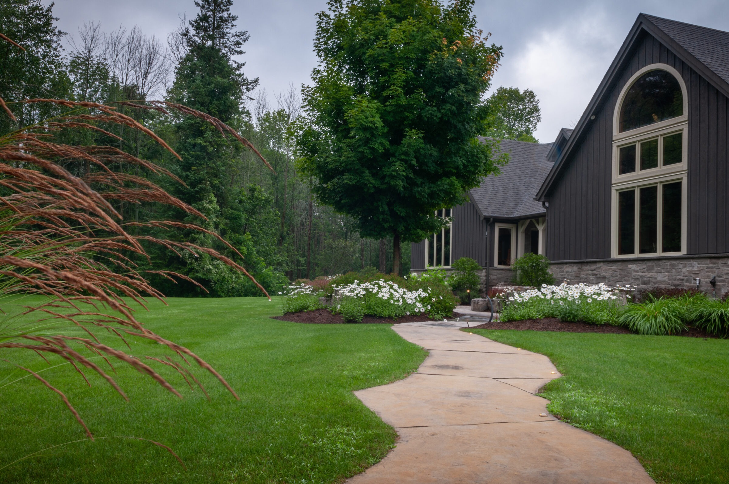 A pathway leads to a large house surrounded by lush green lawn, trees, and white flowers, creating a serene garden scene.