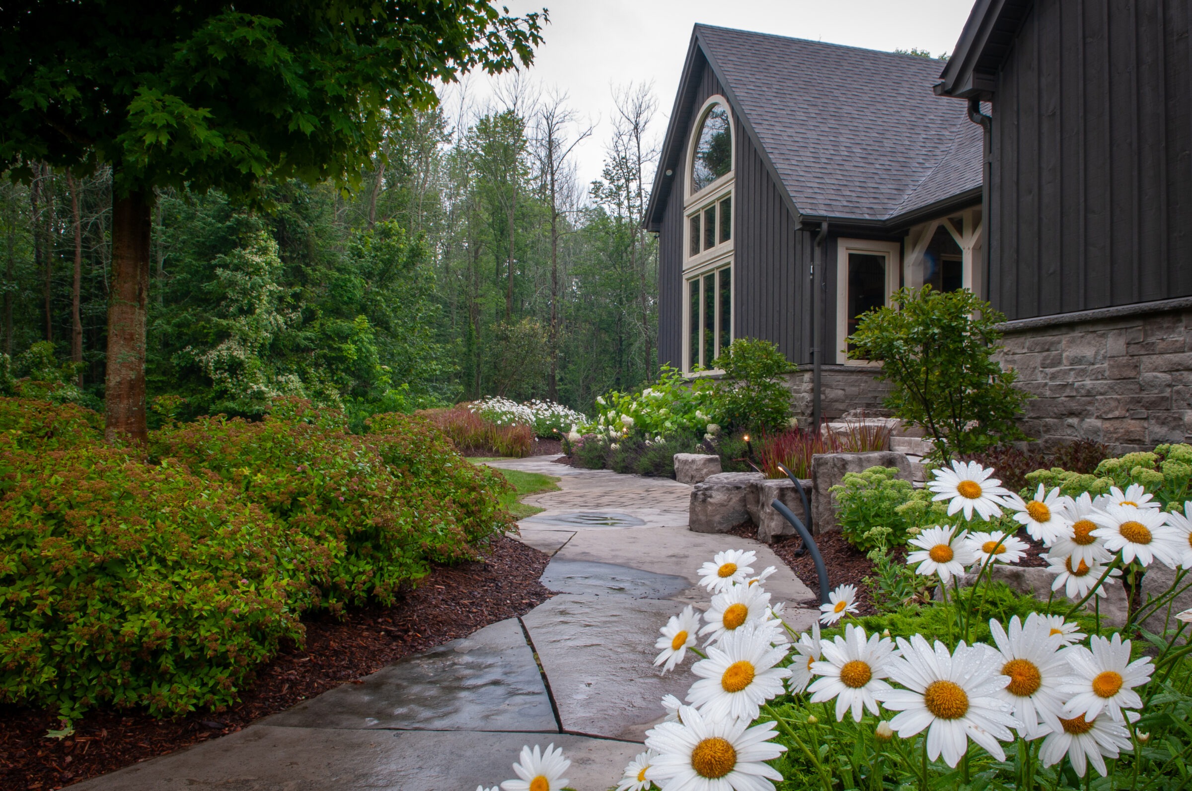 A modern house with stone accents is nestled in lush greenery, featuring a flower-lined pathway and large windows.