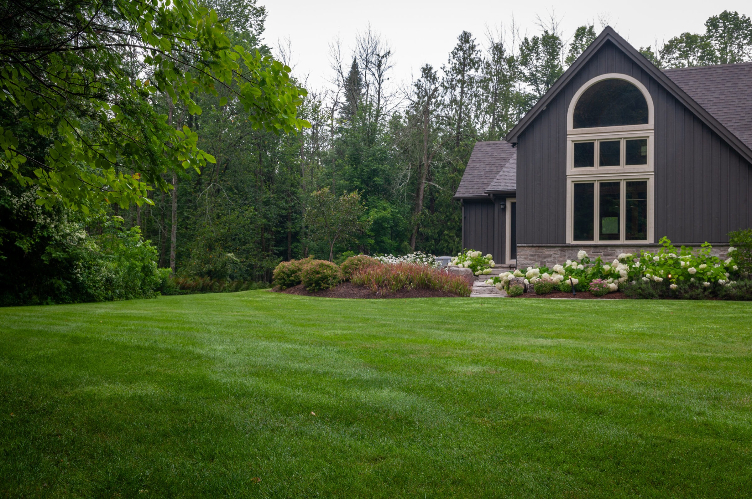 A modern house with a large lawn and garden, surrounded by lush green trees, under an overcast sky.