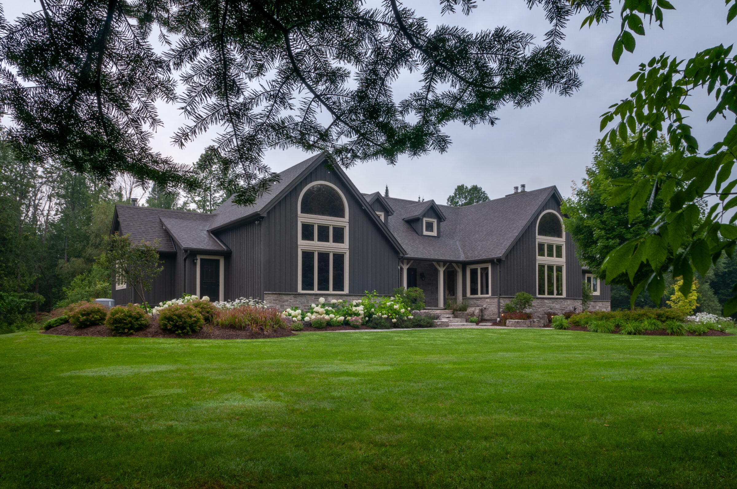 A charming house with large arched windows, surrounded by lush greenery and landscaped gardens, viewed through overhanging tree branches on a cloudy day.