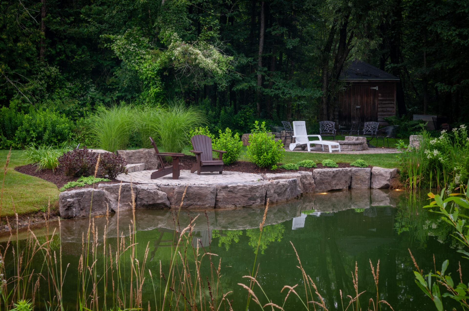 A serene garden scene featuring a small pond, surrounded by lush greenery, stone patio, Adirondack chairs, and a rustic wooden shed.