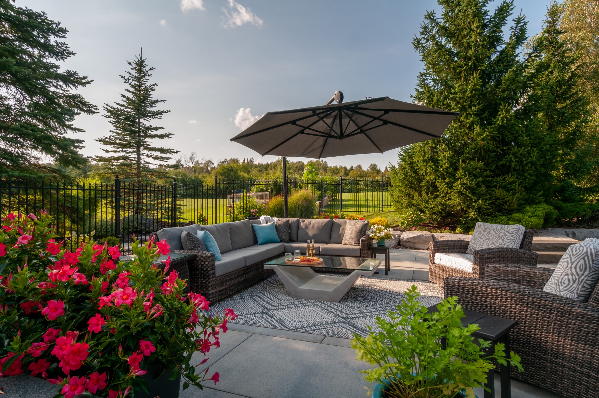 A cozy outdoor patio features wicker furniture, colorful cushions, and vibrant flowers, under a large umbrella, surrounded by lush greenery and a fenced yard.