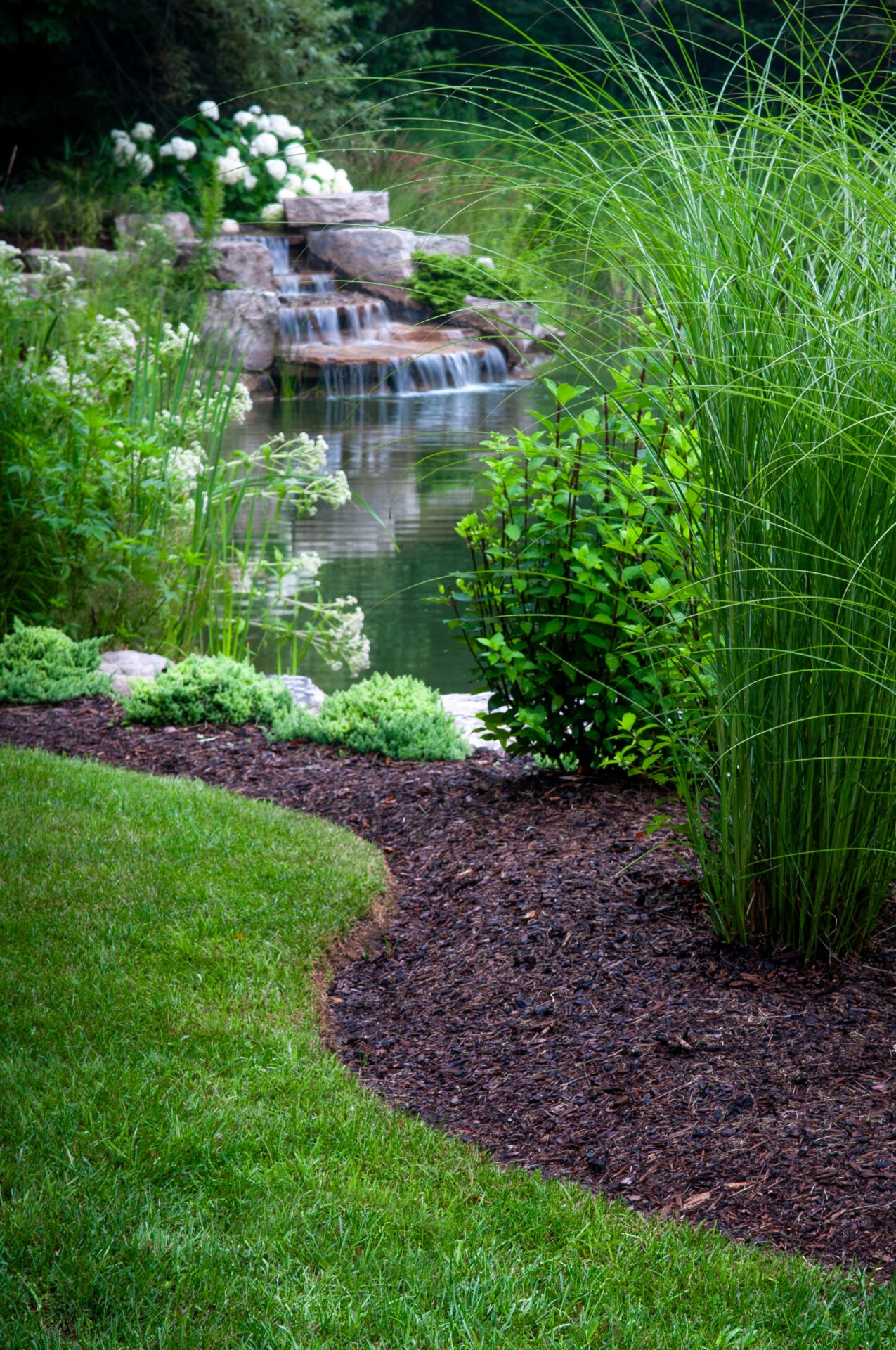 Lush garden scene featuring a small waterfall cascading into a pond, surrounded by vibrant greenery, white flowers, and well-maintained landscaping.