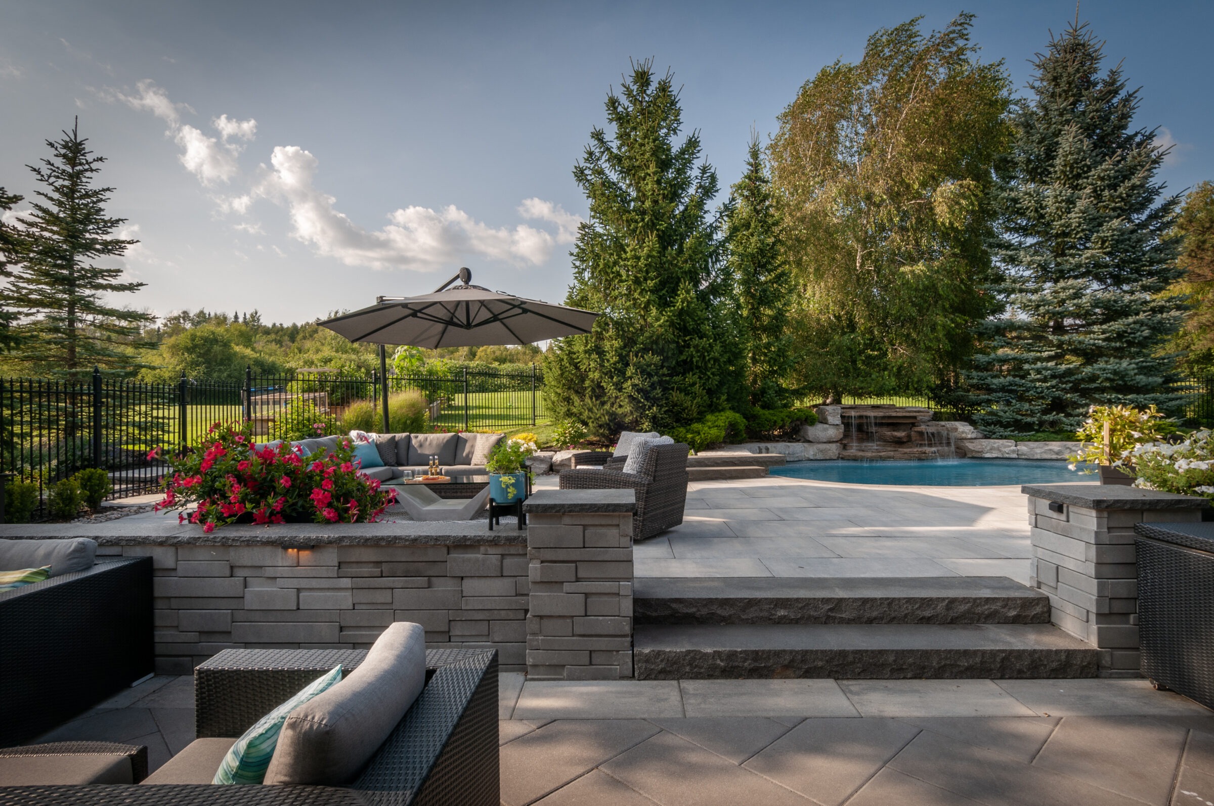 Elegant backyard with pool, patio furniture, and umbrella. Surrounded by lush greenery and trees, creating a serene outdoor retreat under a clear sky.