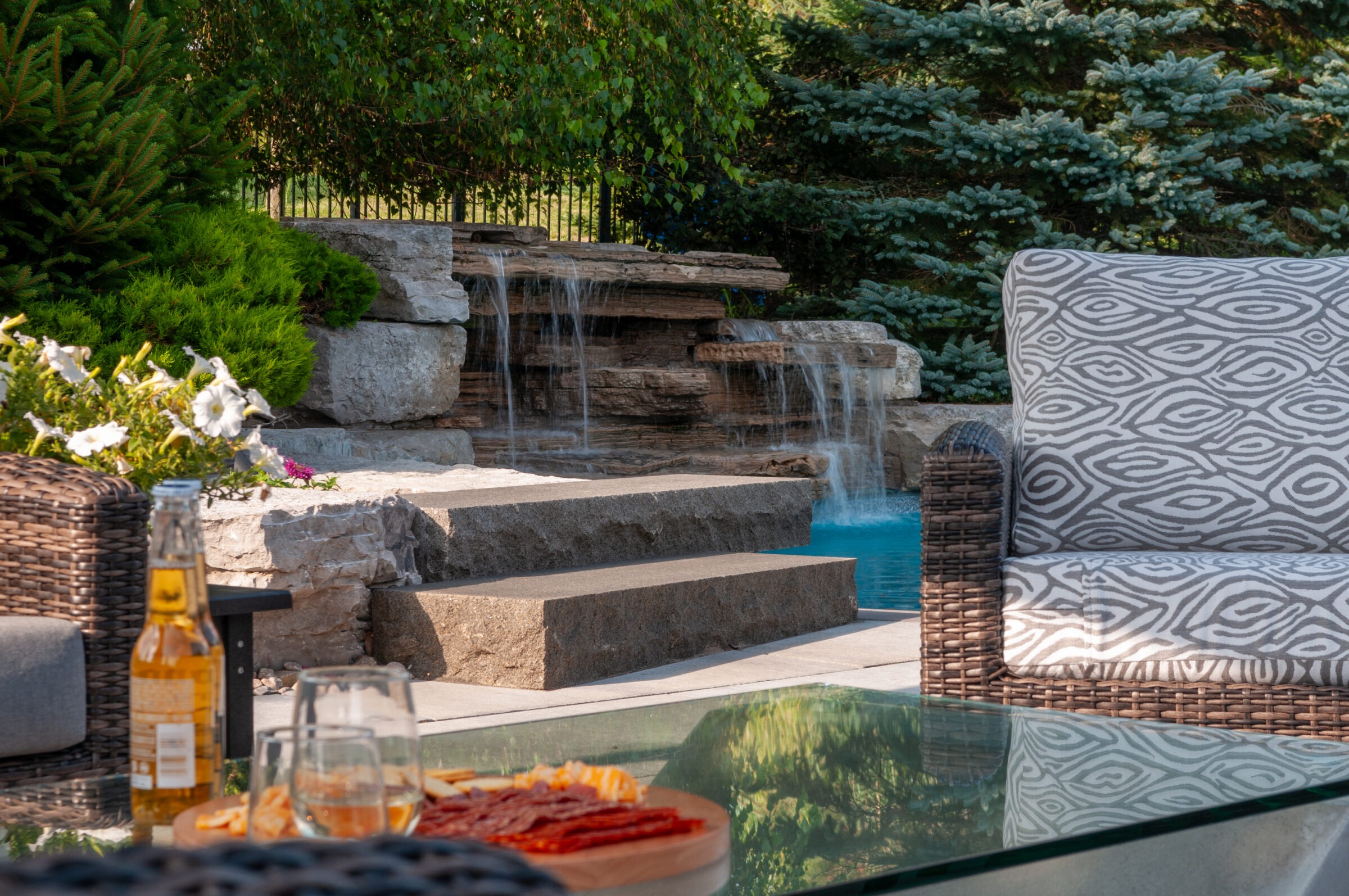 Outdoor seating area with a view of a stone waterfall, surrounded by greenery. Drinks and snacks are on a glass table.
