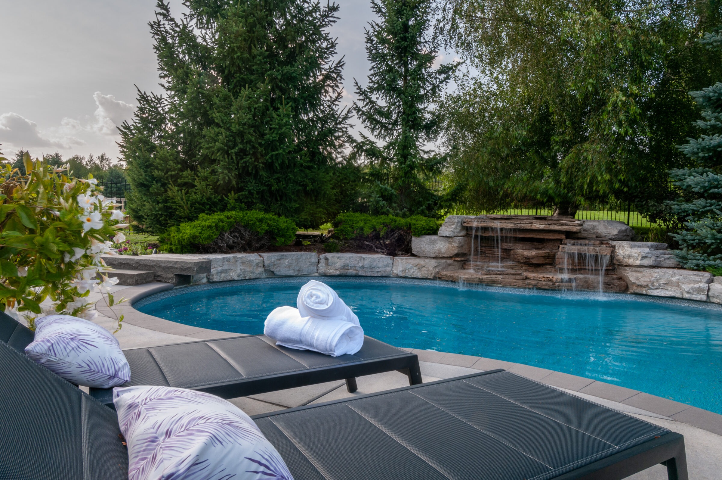 A serene backyard pool with lounge chairs, towels, and cushions beside a small waterfall, surrounded by lush trees and greenery.